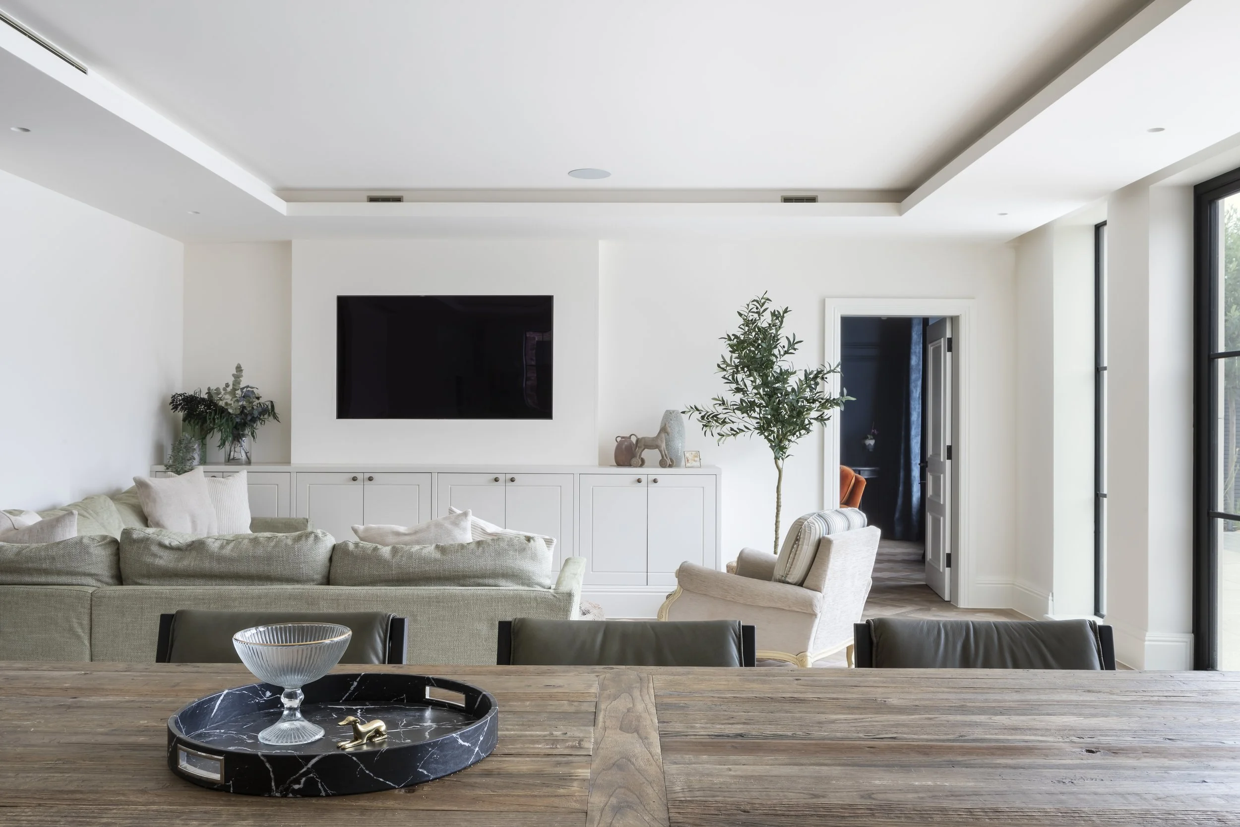 Living room with a large beige sectional sofa, an armchair, a wooden table with a tray holding a glass bowl and small decorative objects. A flat-screen television is mounted above a white cabinet, with decorative items like a small elephant statue an