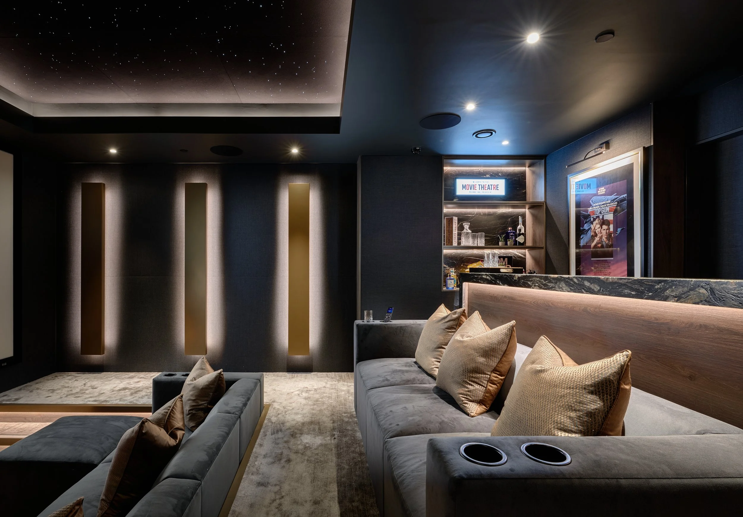 A luxurious home theater room with dark walls, a starry sky ceiling, plush gray sofas with beige cushions, and a bar area with movies and drinks.