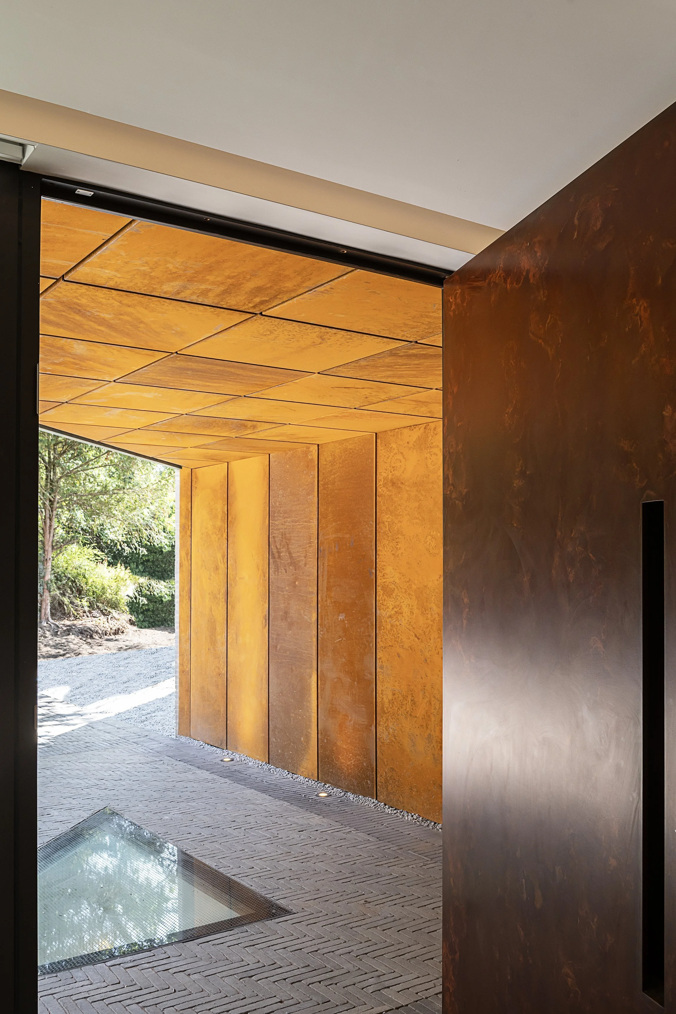 Modern building entrance with rust-colored metal panels on walls and ceiling, brick flooring with a glass window inset, and outdoor greenery visible beyond.