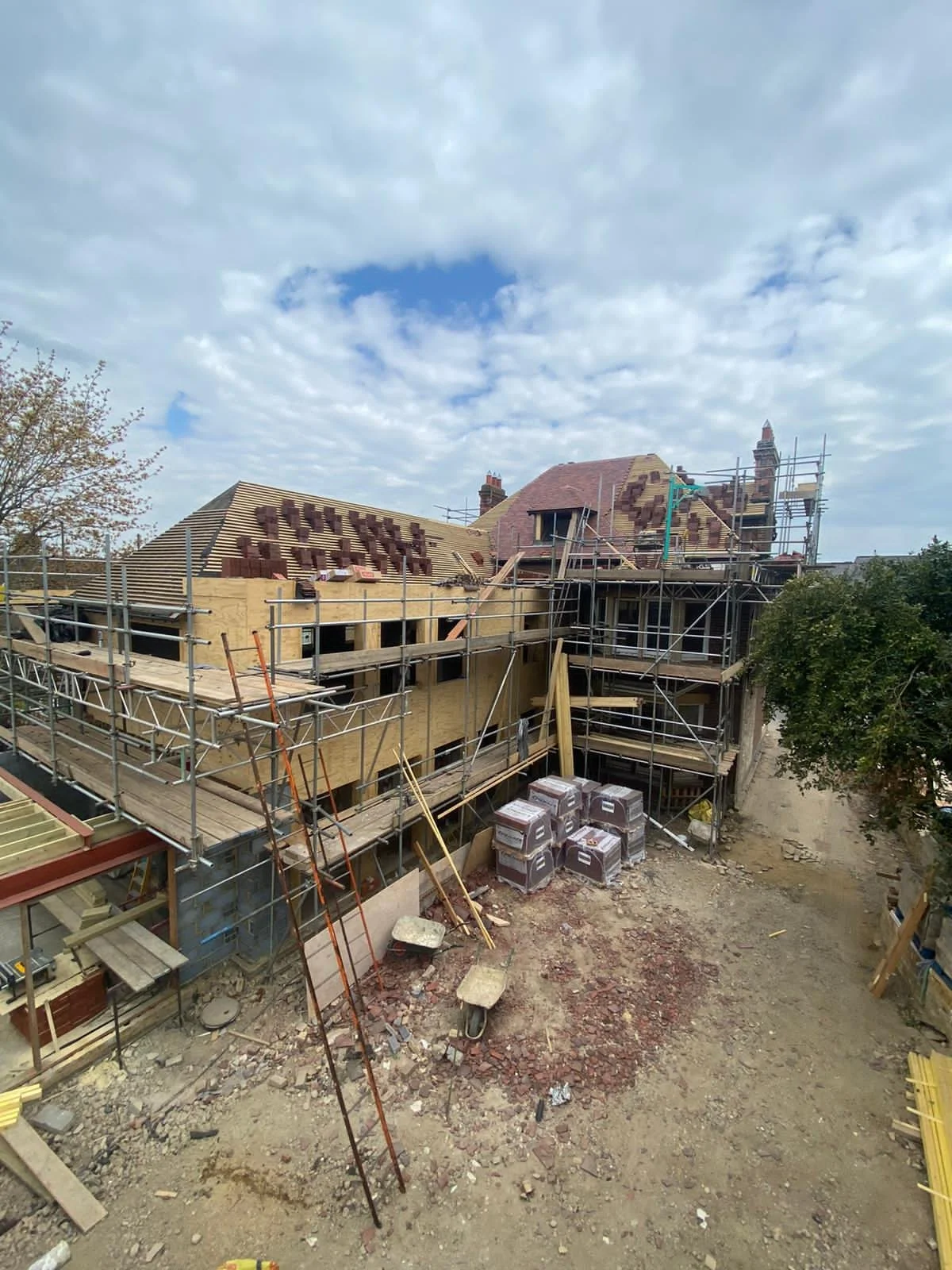 Construction site of a house with scaffolding, roofing materials, and building supplies, under a partly cloudy sky.