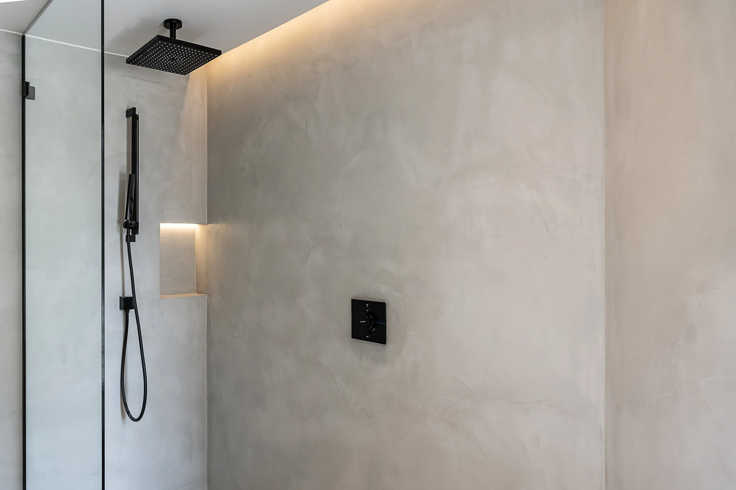 Modern shower with black square rain head, black handheld shower and control on textured beige wall.