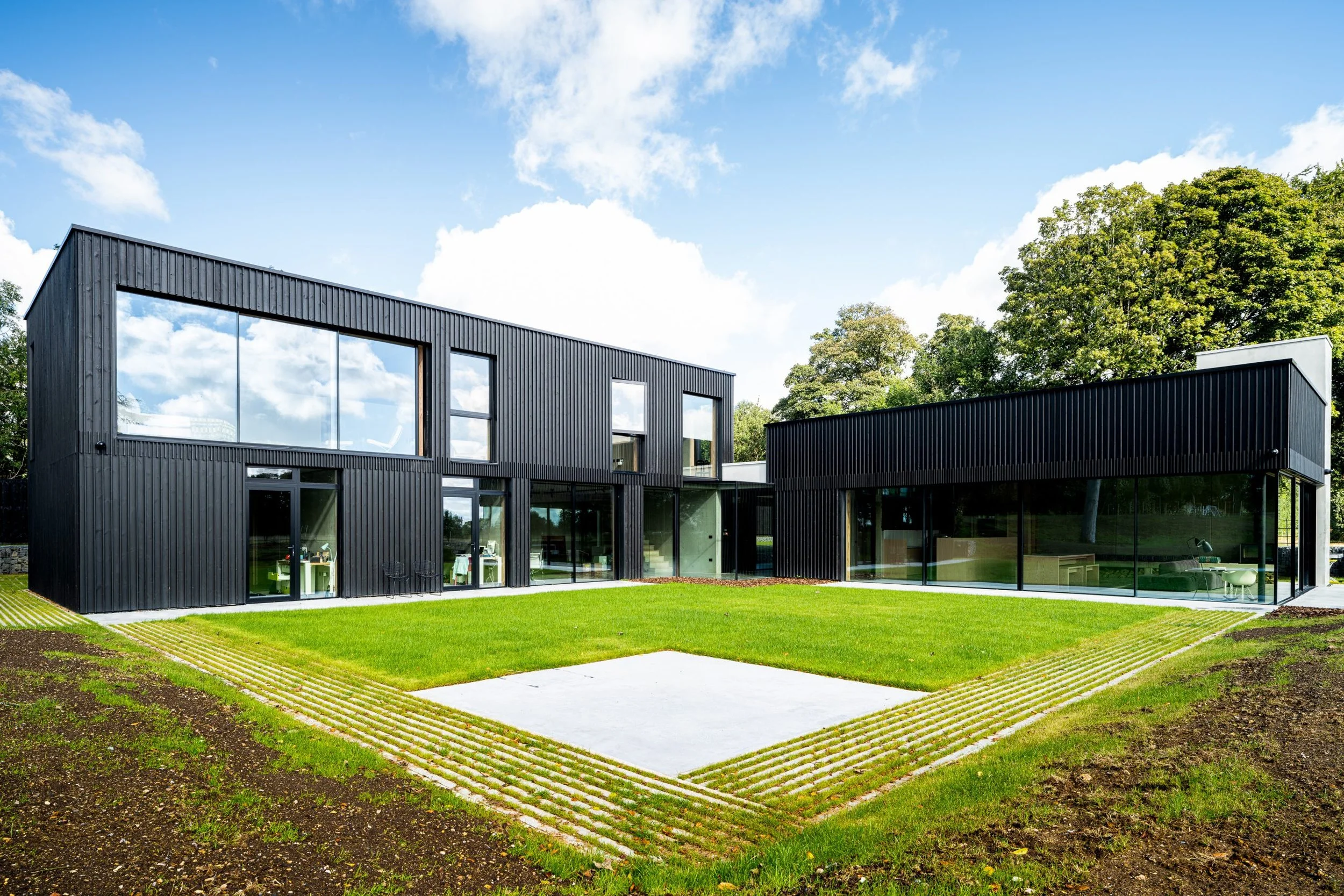 Modern two-story black house with large glass windows, surrounded by a lush green lawn and trees in the background, under a partly cloudy sky.
