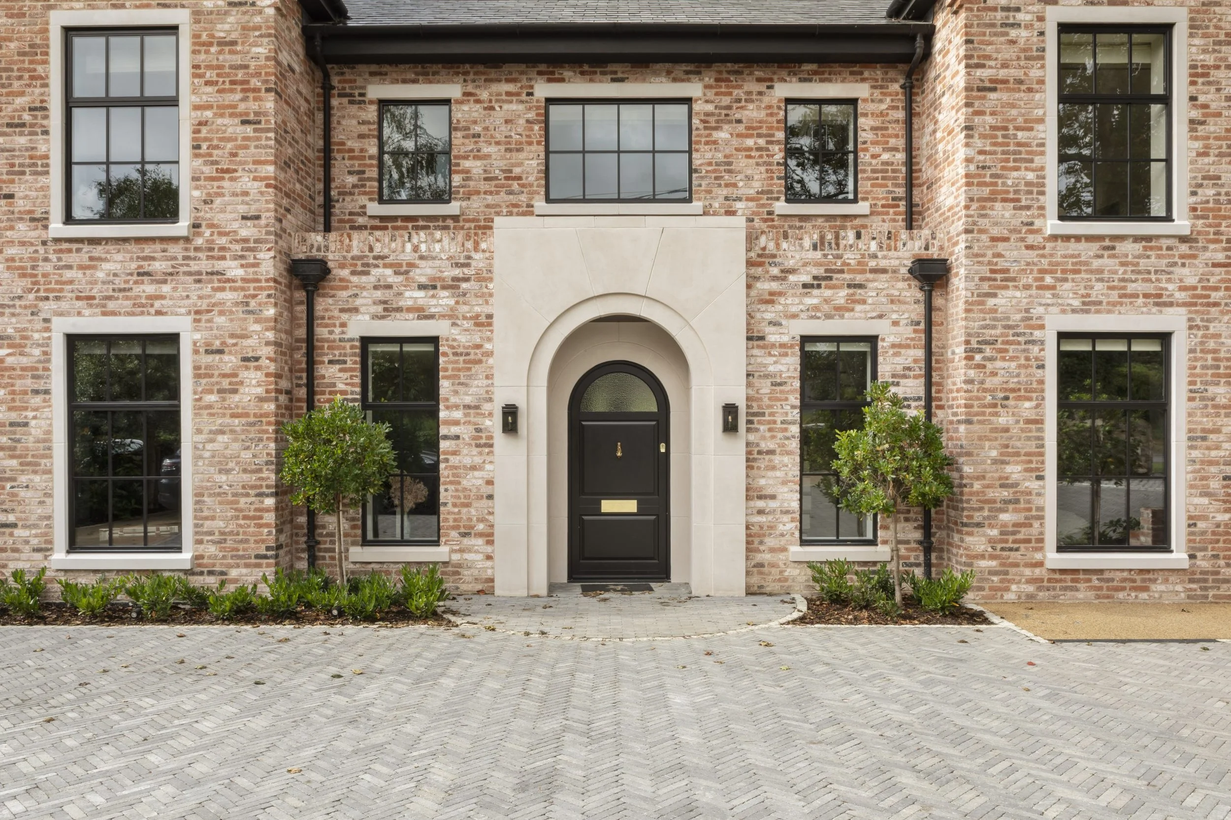 Front view of a brick residential building with black front door, six large windows, two small trees, and a paved driveway.
