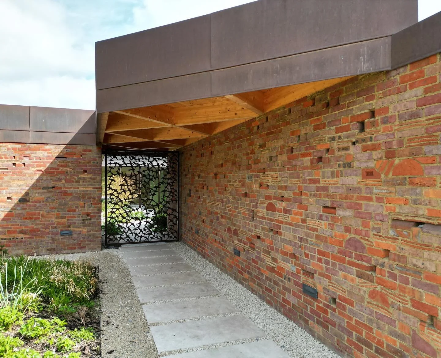 Entryway with brick wall, decorative metal gate, concrete stepping stones, and wooden roof overhang.