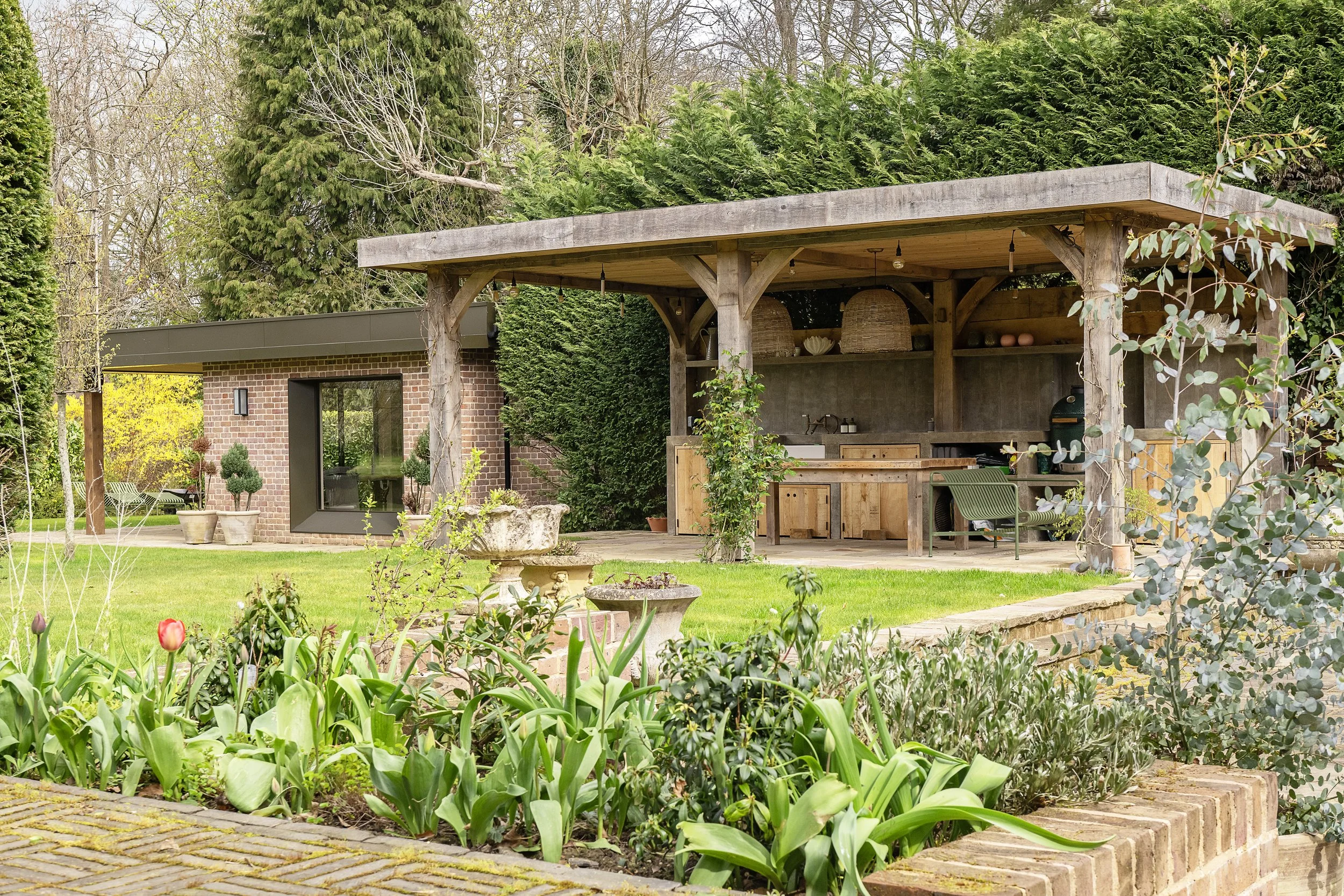 A backyard scene with a wooden outdoor kitchen or bar under a roof, surrounded by green trees and plants, including a garden bed with tulips in the foreground.