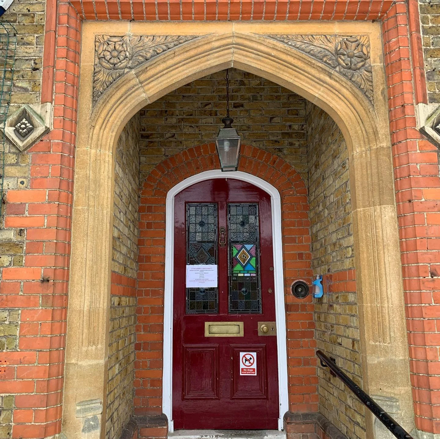 Red front door with stained glass panels, set into a brick wall with decorative stone arch and floral carvings, featuring a hanging lantern, a trash bin, a speaker, and a metallic mail slot, with a 'no dogs' sign on the door.