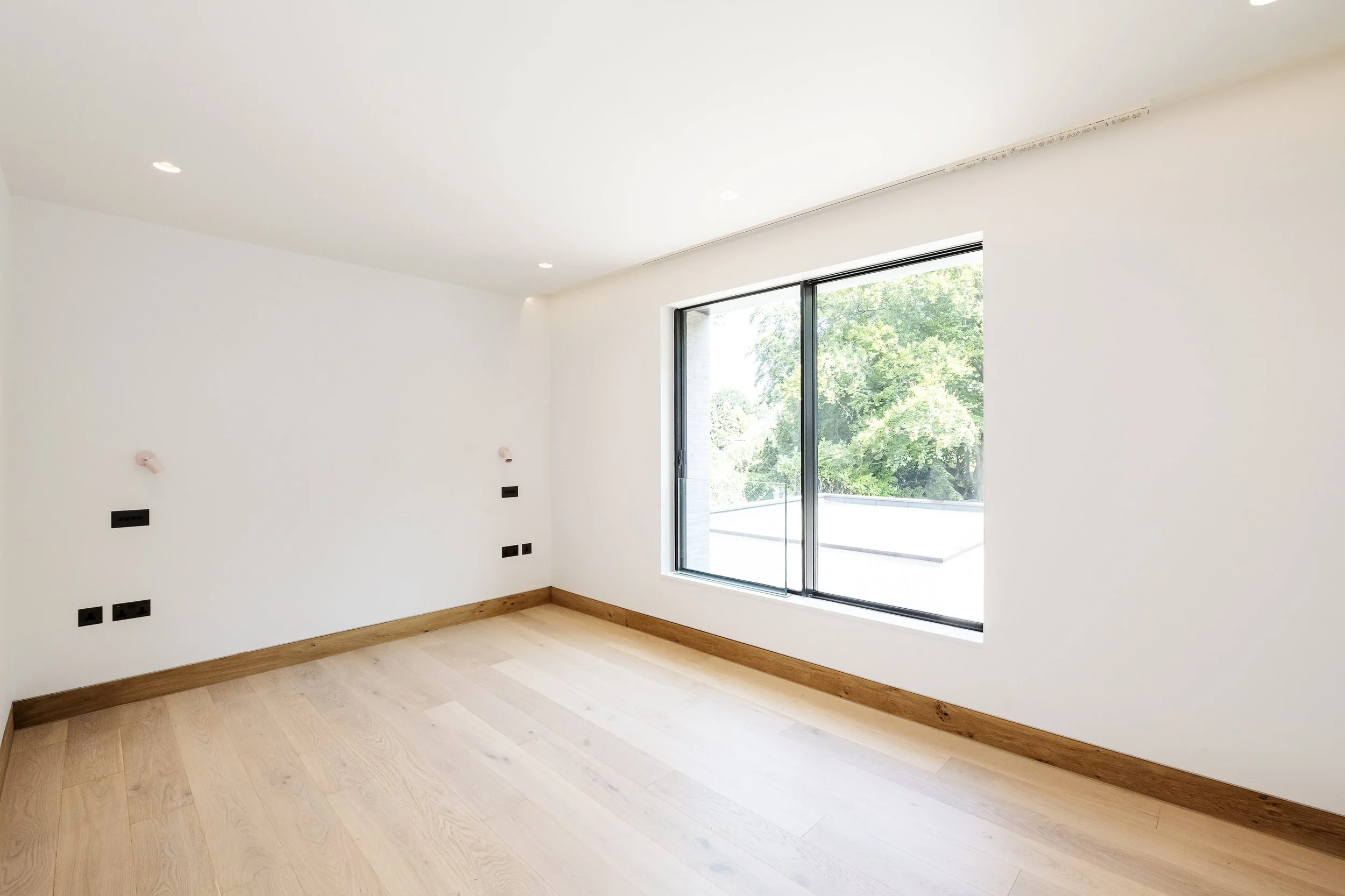 Empty room with white walls, light wooden floor, large window with view of green trees, recess lighting, and electrical outlets.