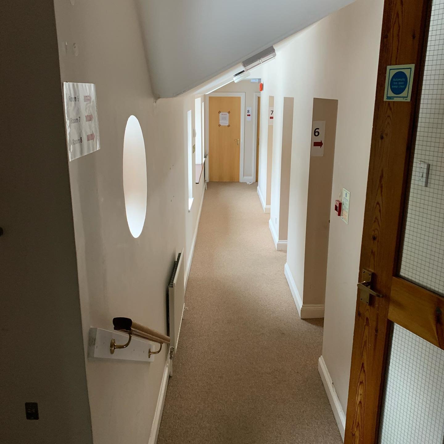 Hallway with beige carpet, white walls, multiple doorways with numbered signs, and a wooden door with a glass panel on the right.