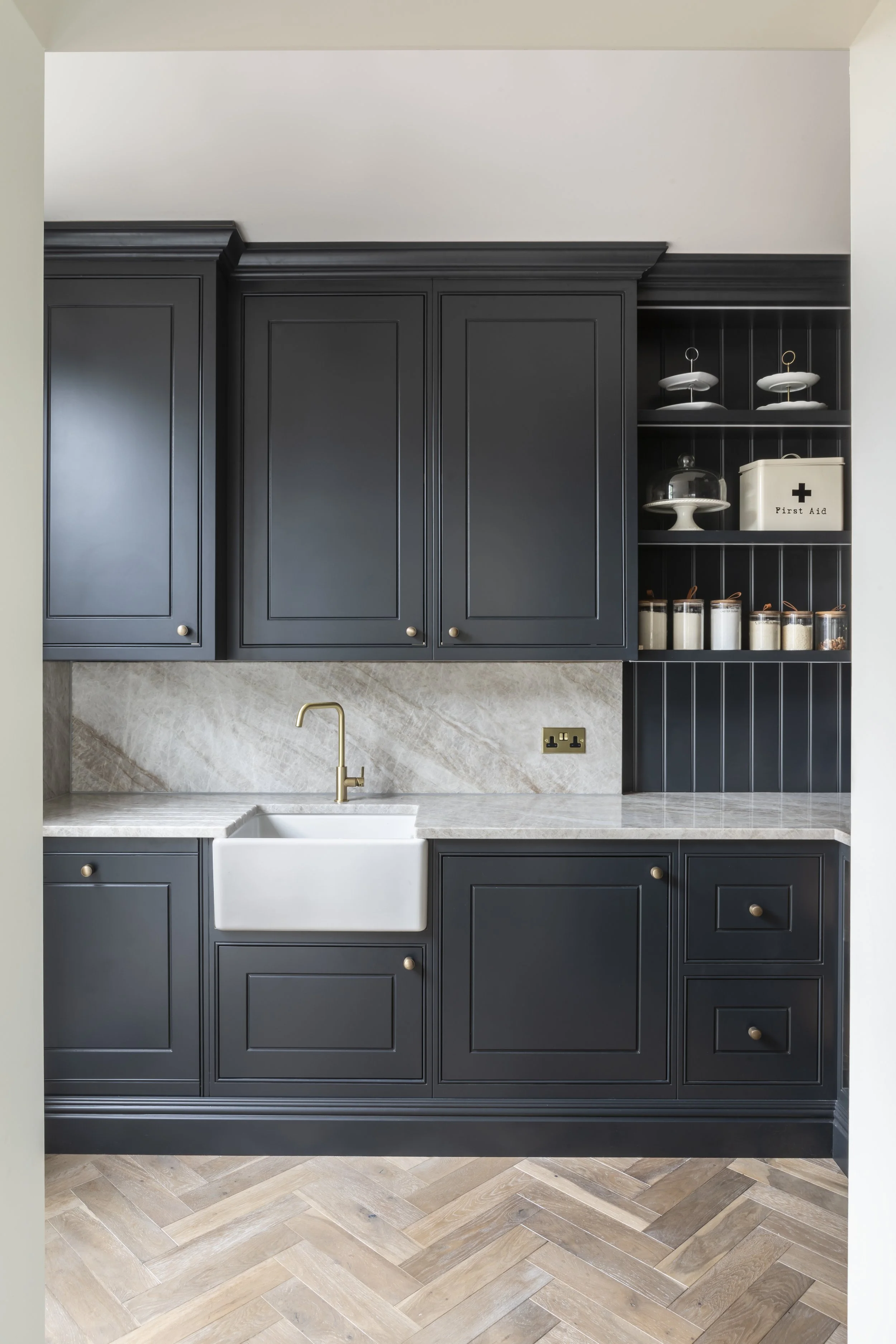 Modern kitchen with dark blue cabinets, marble countertops, and a white farmhouse sink with a gold faucet. Open shelves contain decorative plates, a first aid box, and jars. The floor is patterned wood, and there is a beige wall behind the countertop