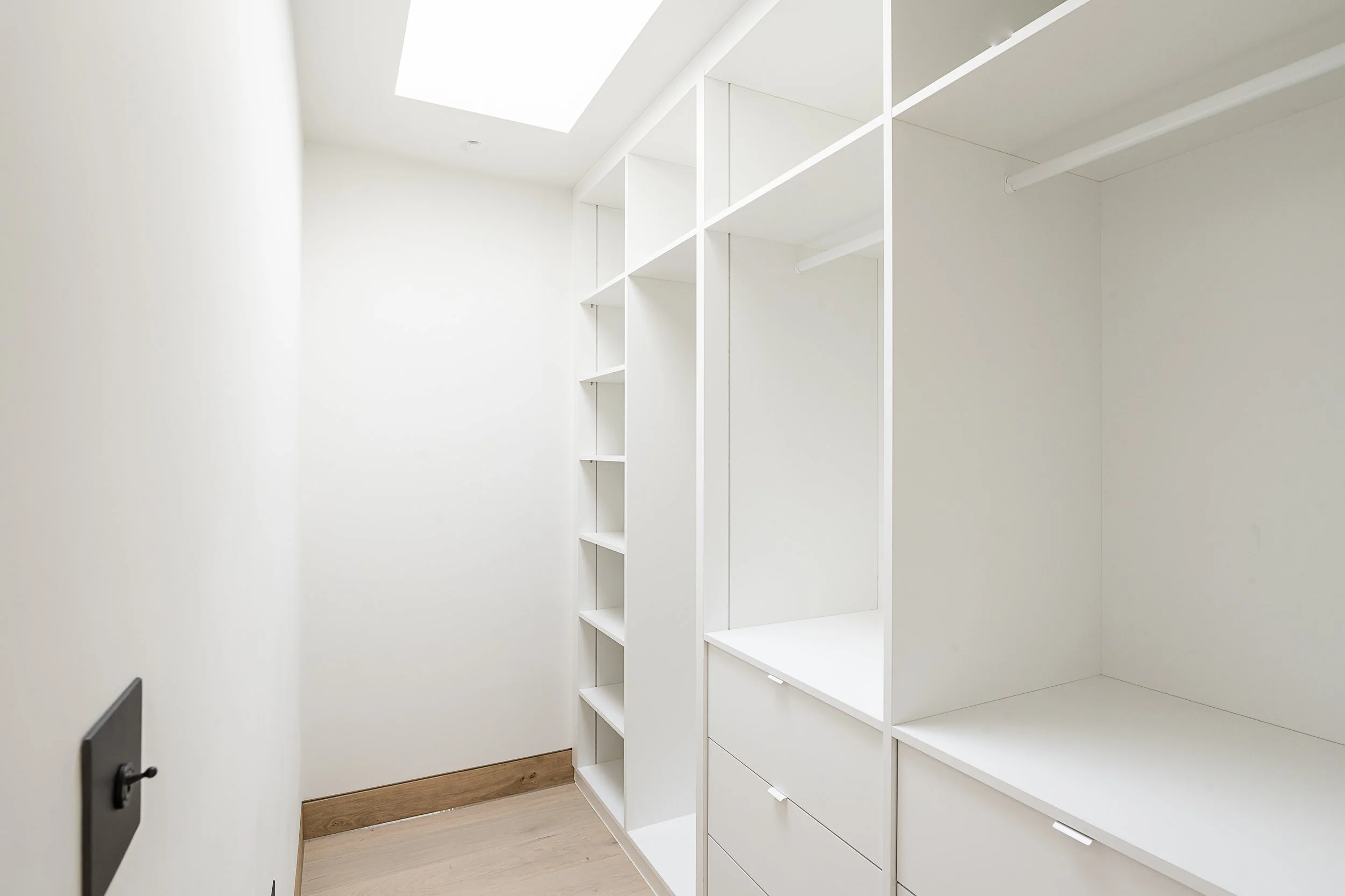 Empty white walk-in closet with built-in shelves and drawers, natural light from skylight.