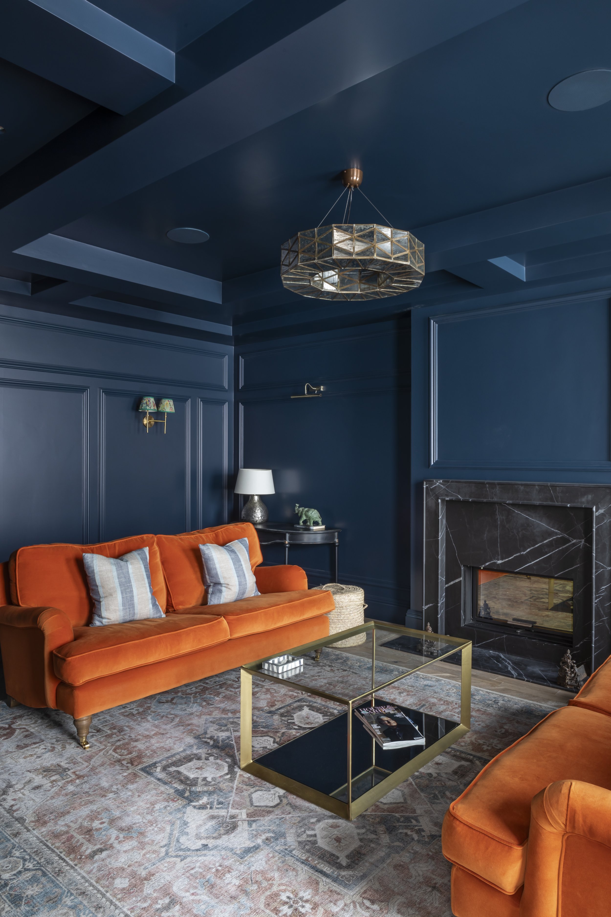 A living room with blue walls and ceiling, featuring an orange velvet sofa with striped pillows, a marble fireplace, a glass coffee table with a magazine, a black side table with a white lamp, and a decorative brass-and-glass chandelier.