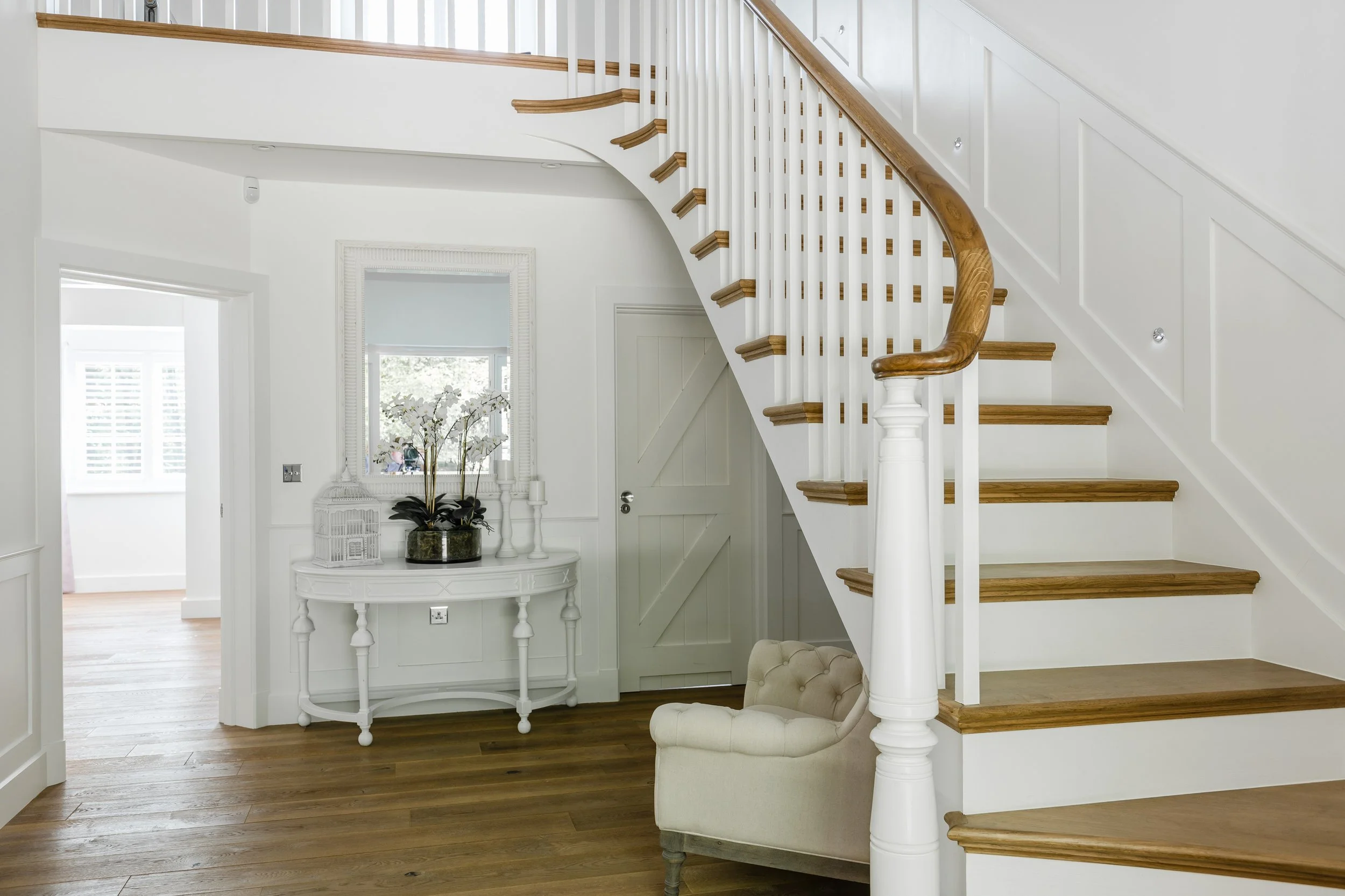 Interior view of a home with hardwood flooring, a staircase with wooden steps and white walls, a white table with plant and candle decorations, and a beige tufted armchair.