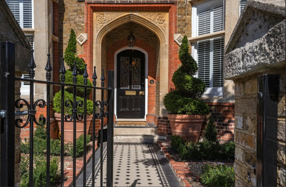 The entrance of a brick house with a black front door, decorative archway, potted topiary bushes, and a black iron gate and fence.