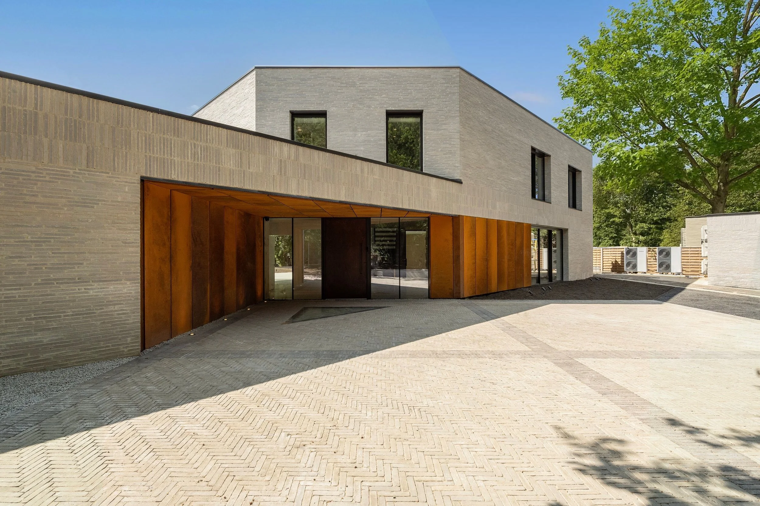 Modern two-story building with gray brick facade, large windows, and a wooden covered entrance area, surrounded by a paved driveway and greenery.