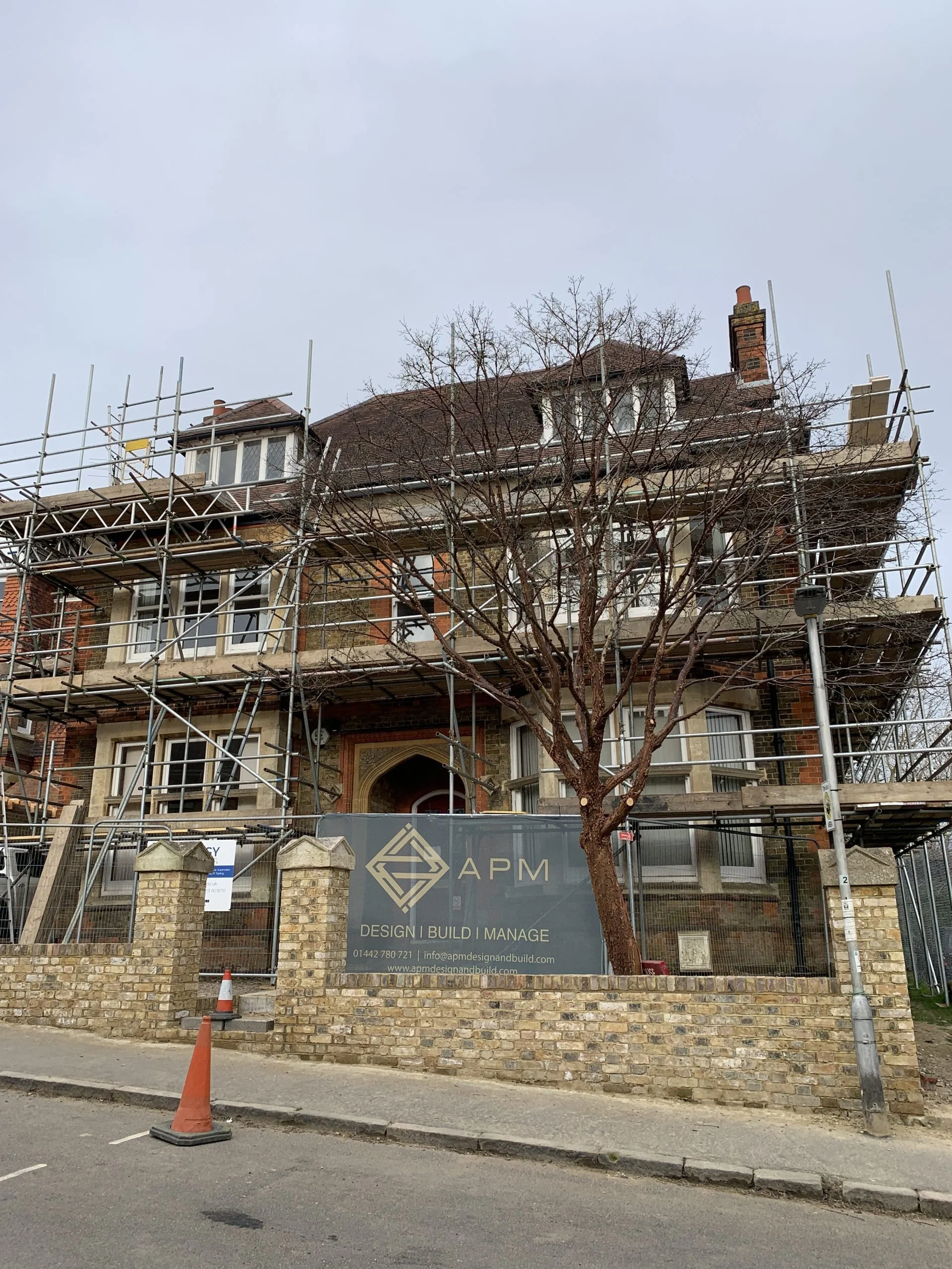 A multi-story brick and stone house under renovation, surrounded by metal scaffolding, with a sign for APM design and build in front of a brick wall and a leafless tree.