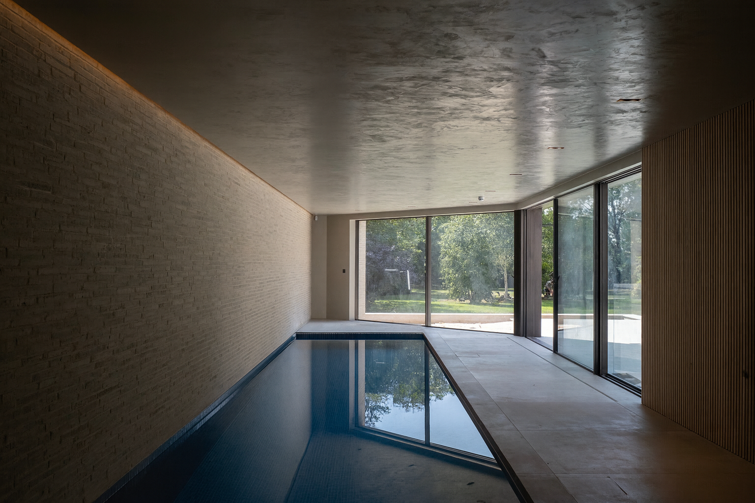 Indoor swimming pool with large glass windows showing green trees outside, modern minimalist design with wooden and brick walls.