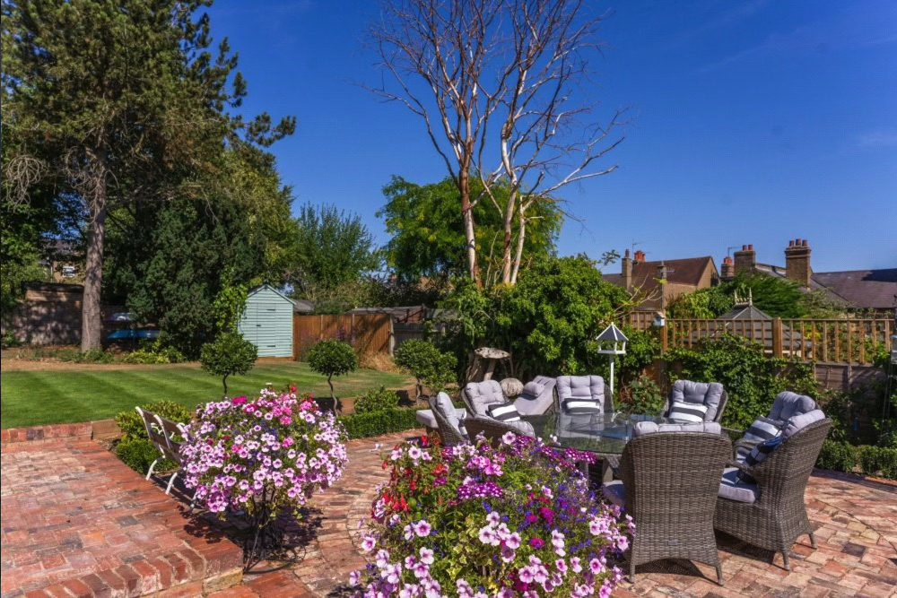 Backyard patio with potted flowers, patio furniture and lush green garden under a blue sky.