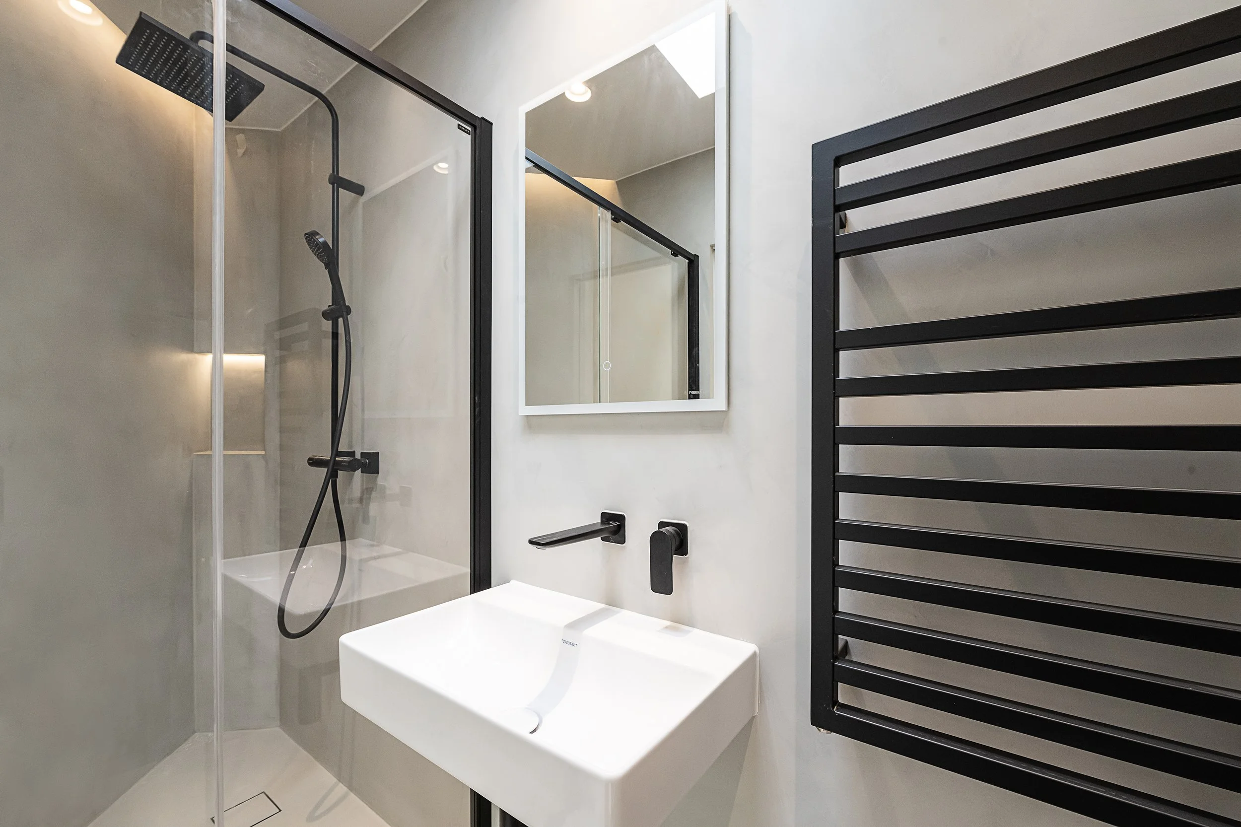 Contemporary bathroom with glass-enclosed shower, black fixtures, wall-mounted white sink, mirror, and black heated towel rack.