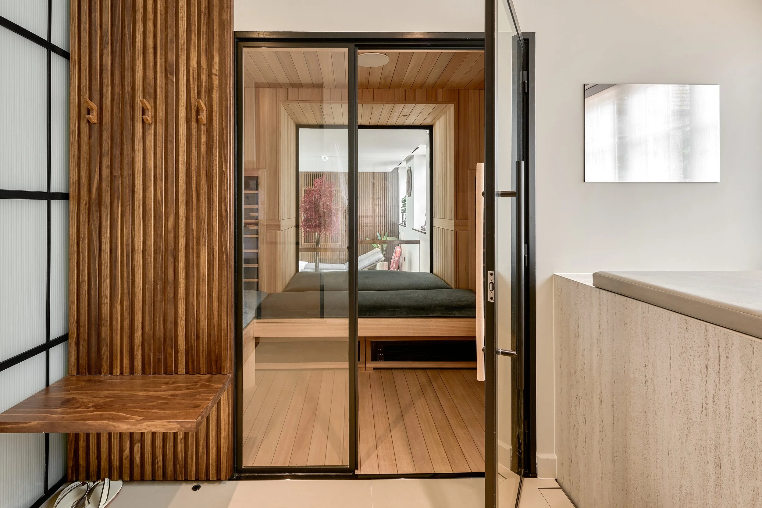 A modern interior view of a bedroom seen through glass sliding doors, with wood-paneled walls, a bed, and decorative plants.