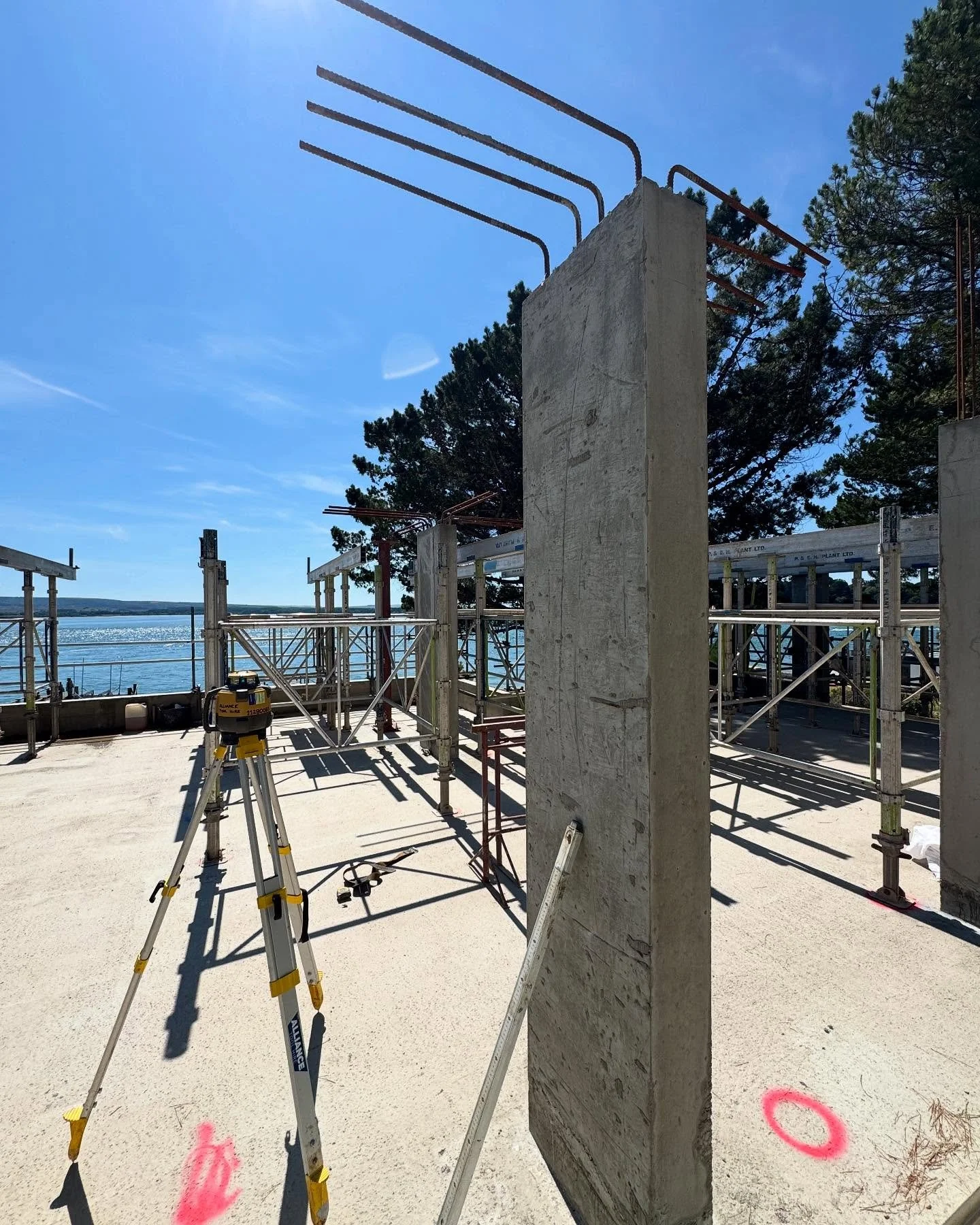 Construction site near water with concrete wall, scaffolding, surveying equipment, and trees in the background under clear blue sky.