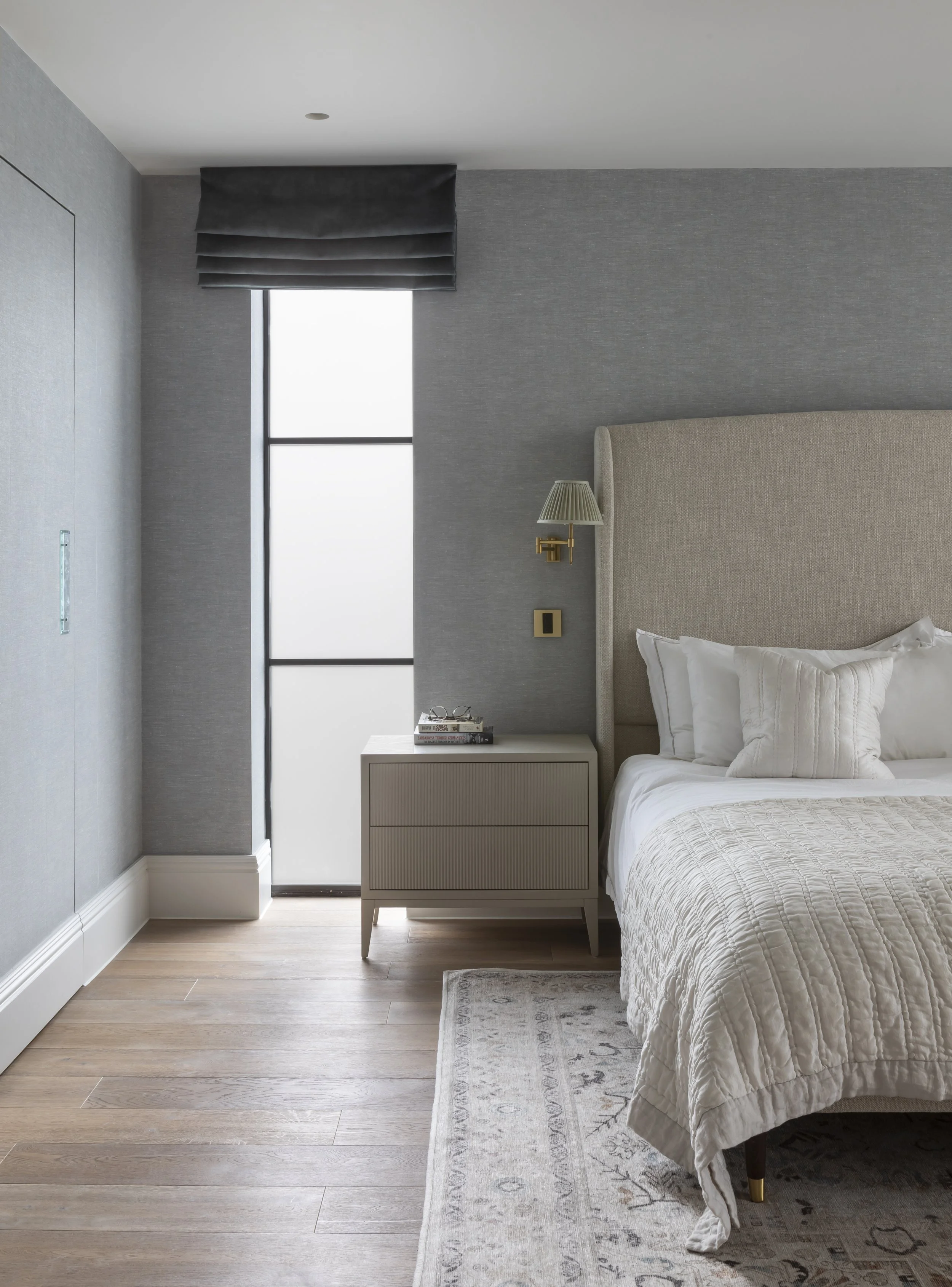 A modern bedroom with a light beige upholstered bed, white bedding, a gray nightstand, a small lamp, and a window with frosted glass and a black valance. The room has gray walls, light wooden flooring, and a patterned area rug.
