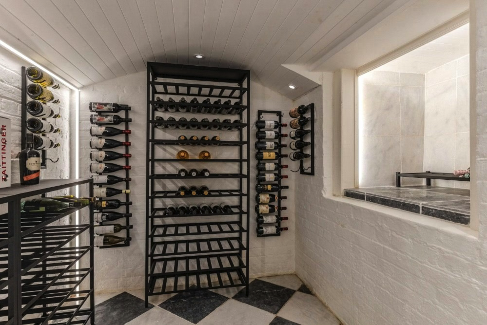 Wine cellar with black metal wine racks holding multiple bottles of wine, situated against white brick walls and a white wooden ceiling.