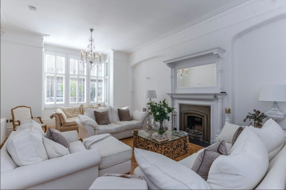 Living room with white sofas, a chandelier, large window with shutters, and a fireplace with a mirror above it.