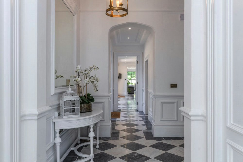 Bright, white hallway with checkered black and white marble flooring, decorative molding, and a hanging light fixture. There is a small white table with plants and a cage decoration on it. Further down the hall, a room with large windows and a view o