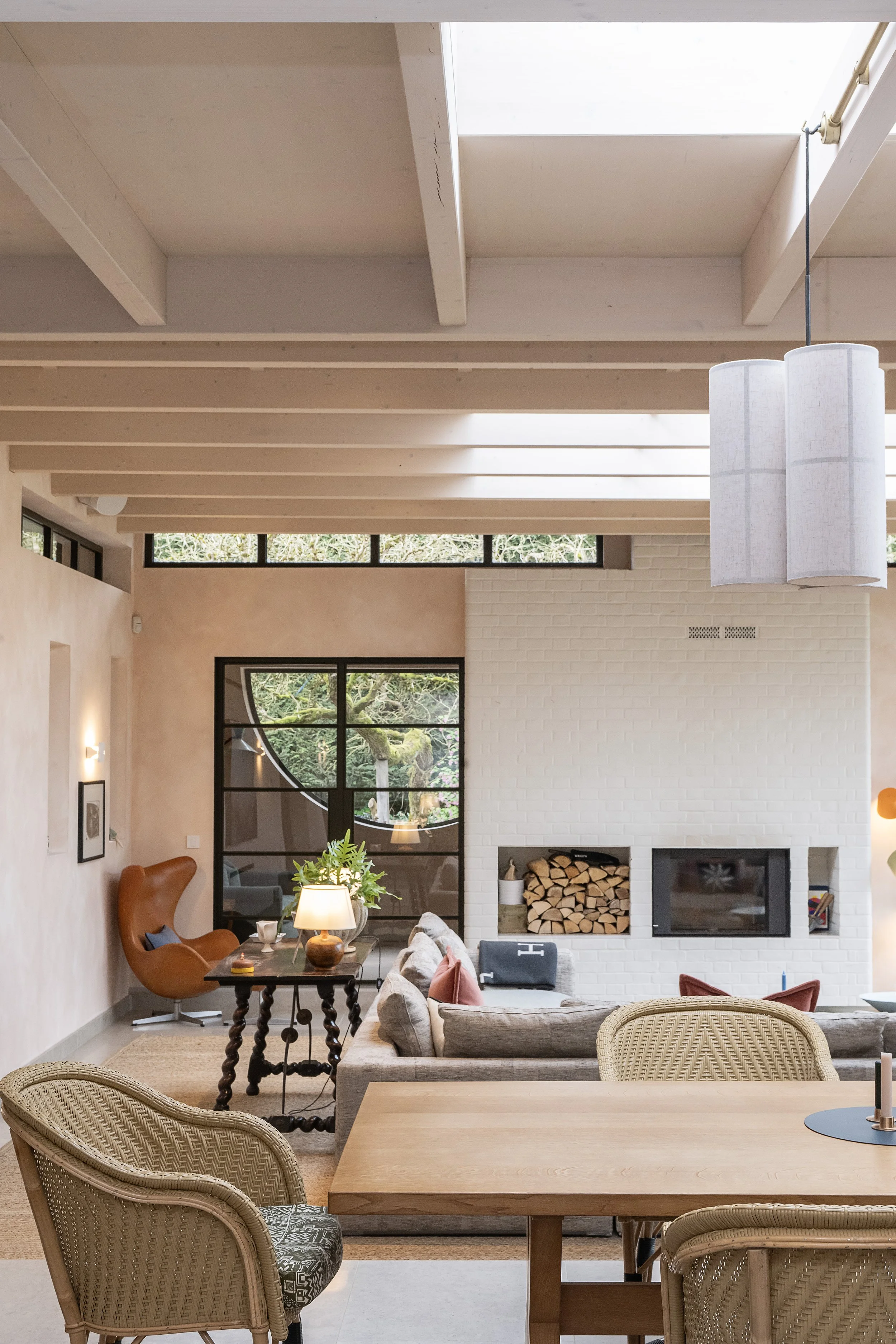 Living room with white brick fireplace, wood logs, beige sofa, wicker chairs, wooden dining table, pendant lights, and large window with an arched design showing outdoor greenery.
