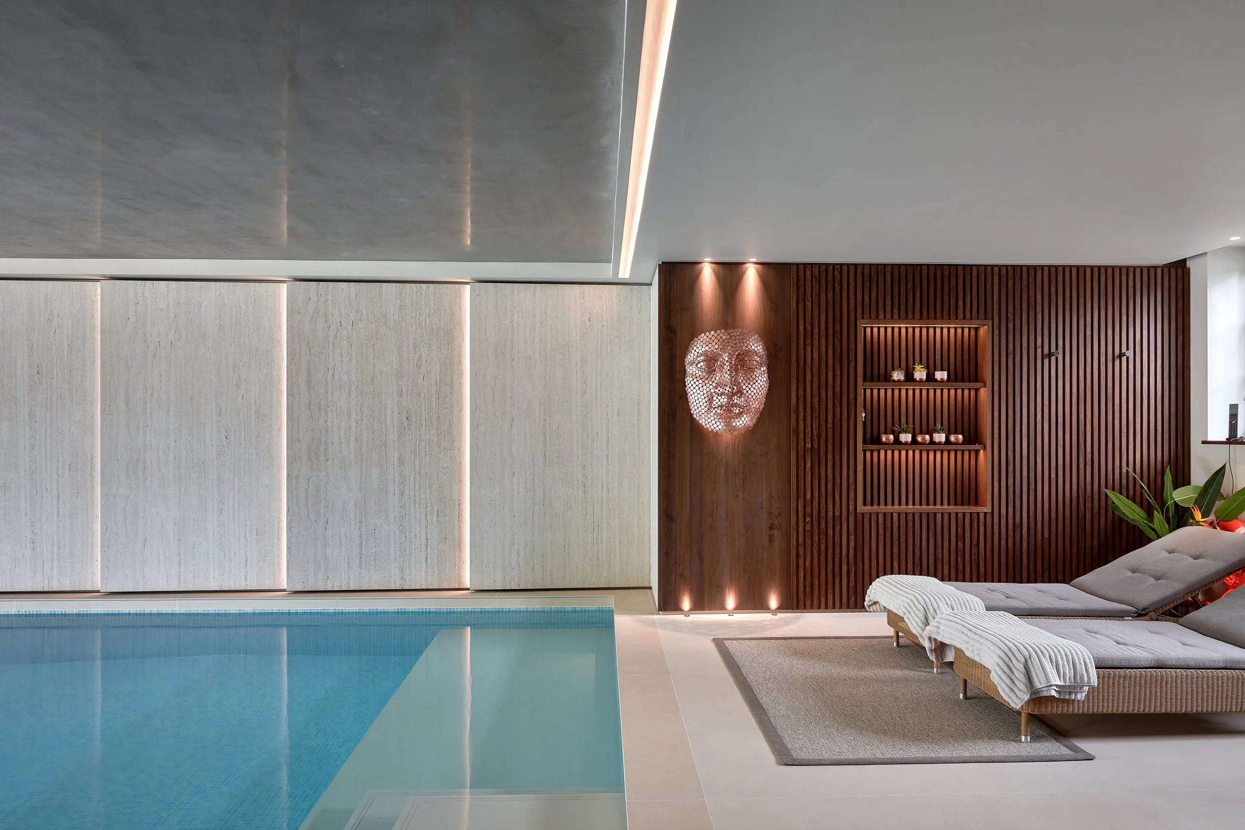 Modern indoor swimming pool area with lounge chairs, wooden wall decor, and a decorative face sculpture with lighting.