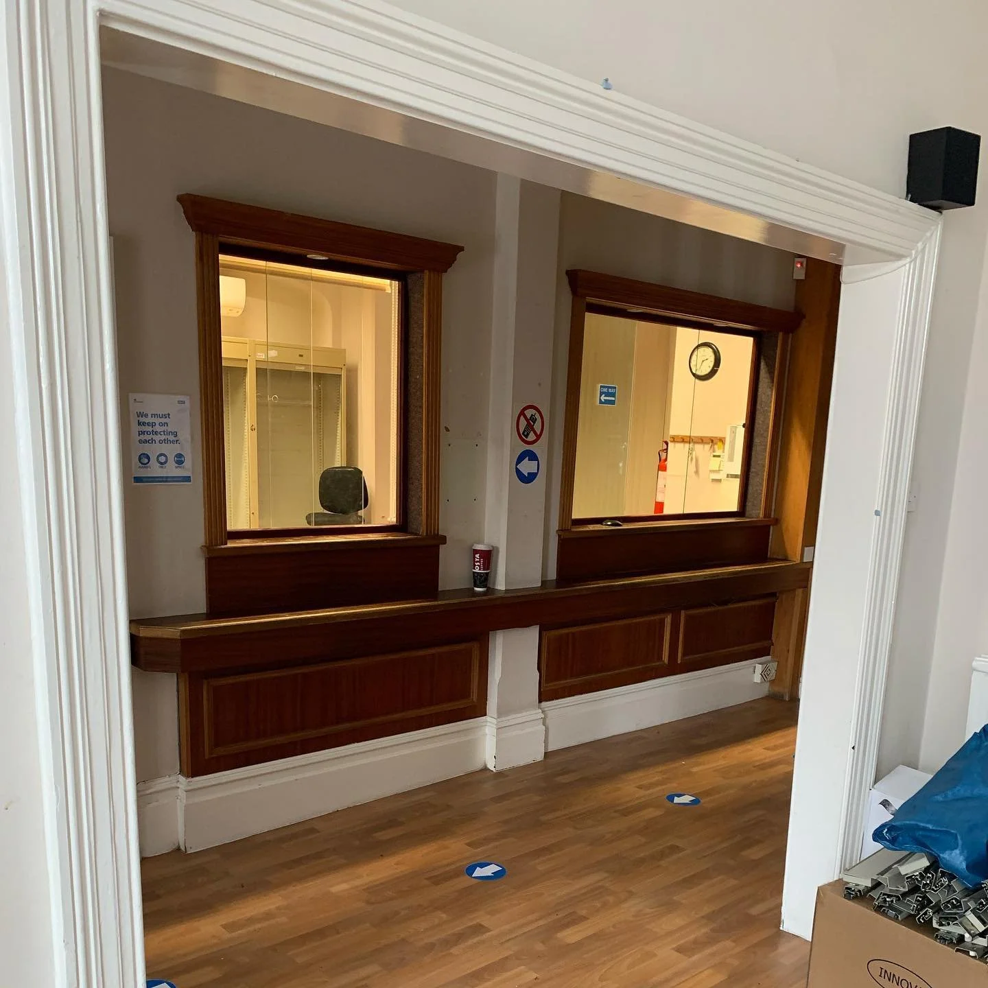 Interior view of a reception window area with two service counters, one of which has a coffee cup and a fire alarm. There are directional arrows on the floor and signs on the walls indicating COVID-19 safety measures and directions.