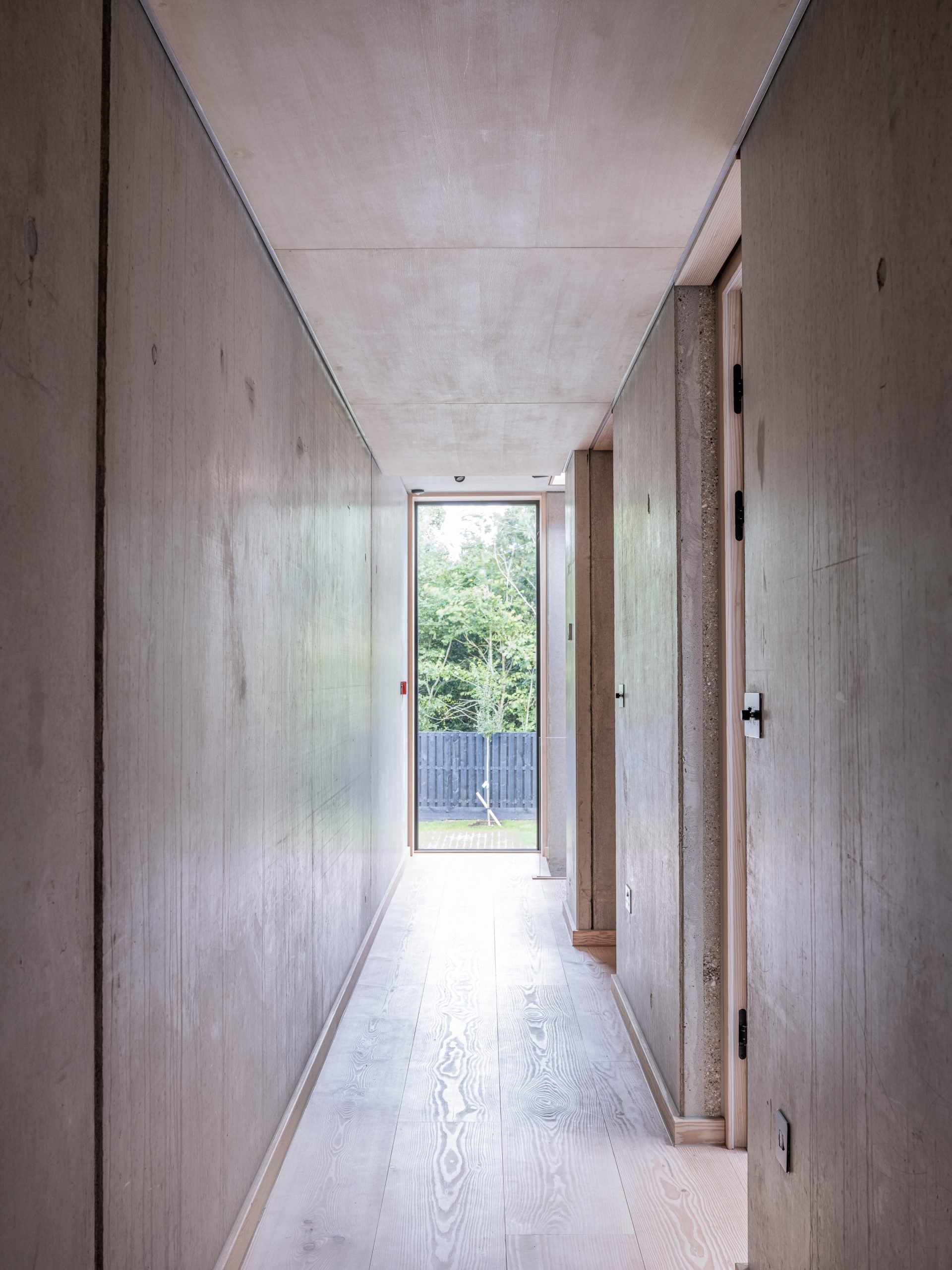 A narrow hallway with light-colored wooden floors and walls, leading to a glass door at the end with trees visible outside.