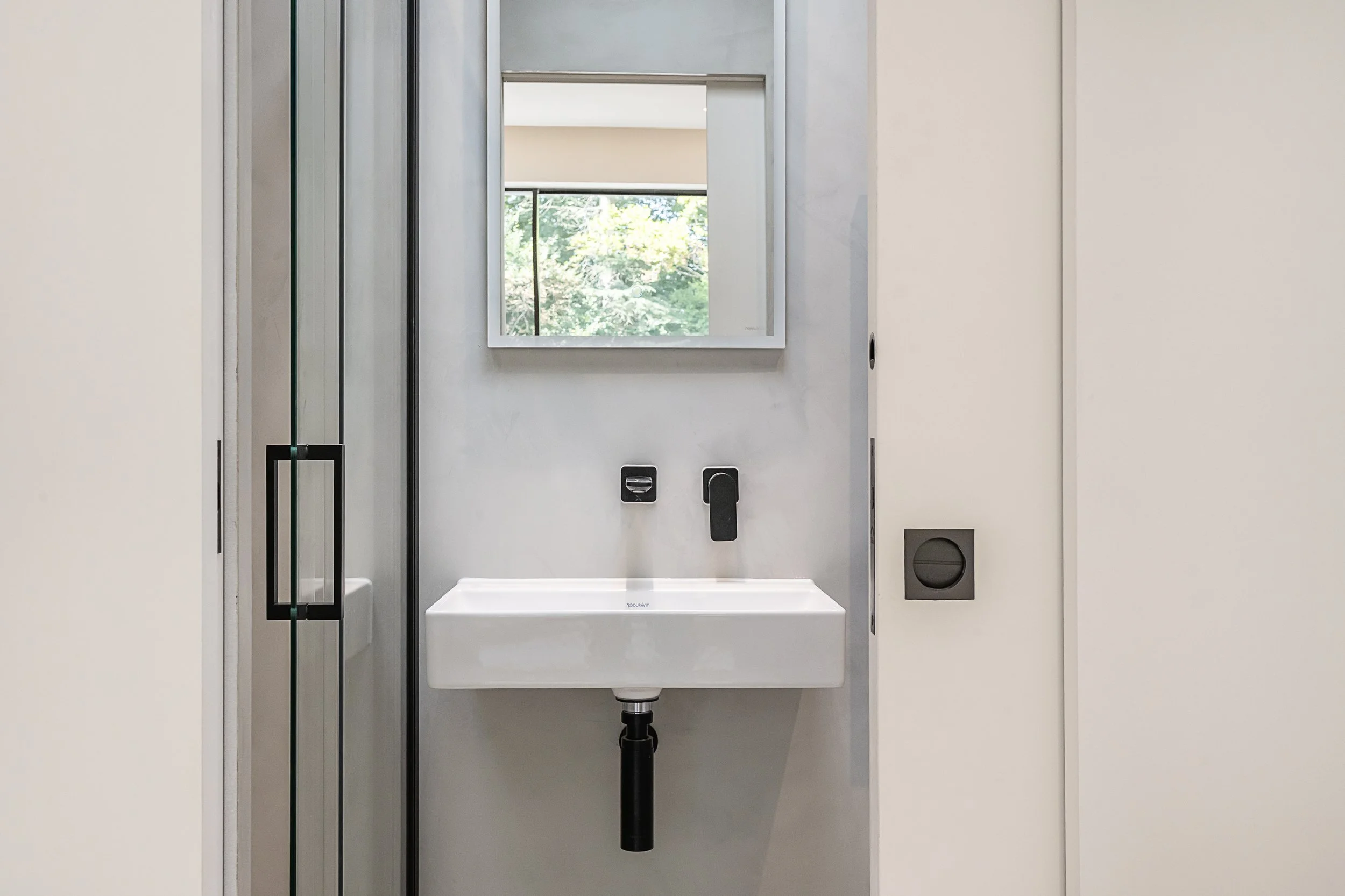 Small white wall-mounted sink beneath a mirror, with black faucet and plumbing, in a modern bathroom with a window showing green trees outside.