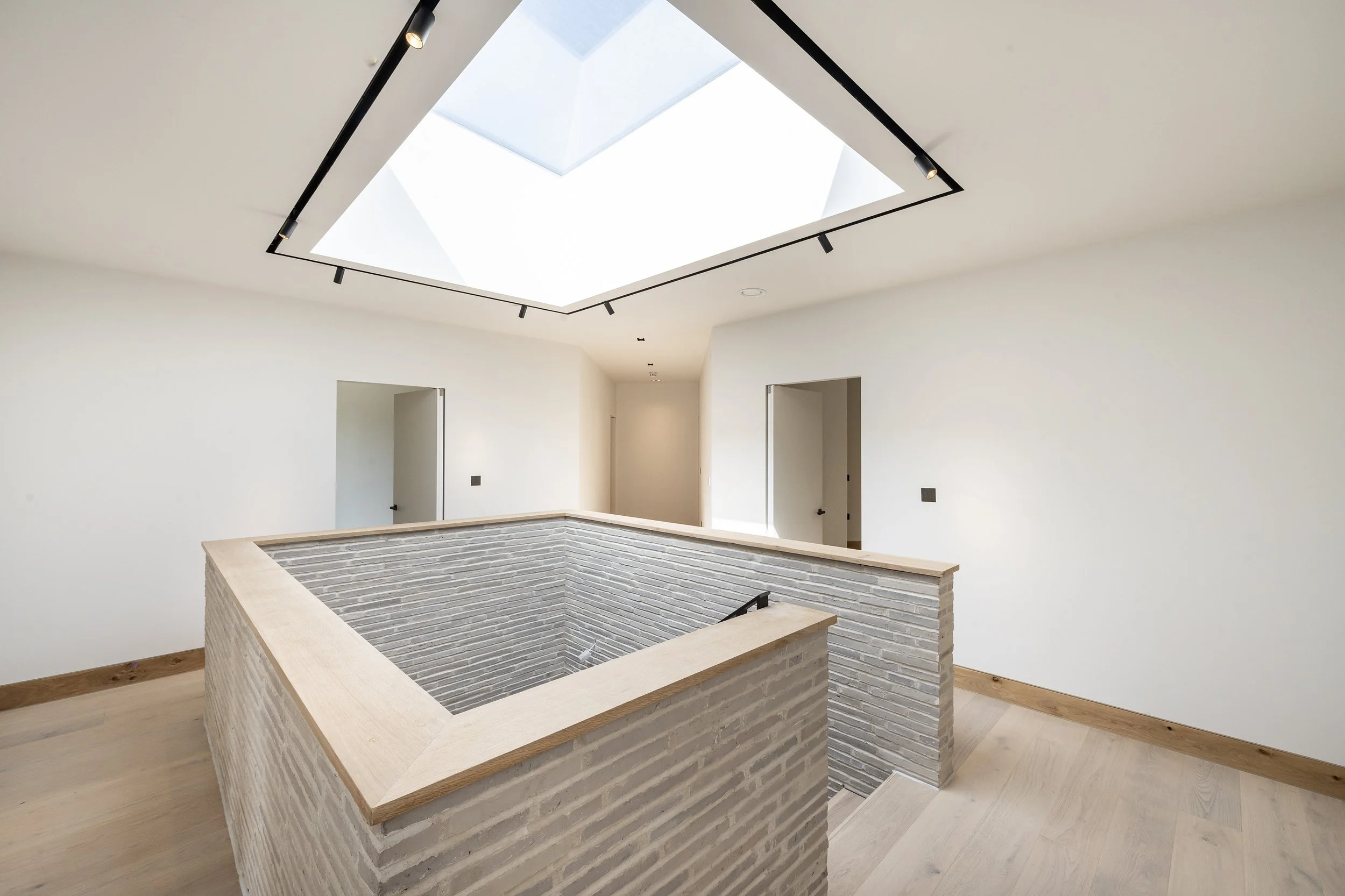 Empty modern interior room with white walls, wooden flooring, an open skylight ceiling, and a brick-walled opening.