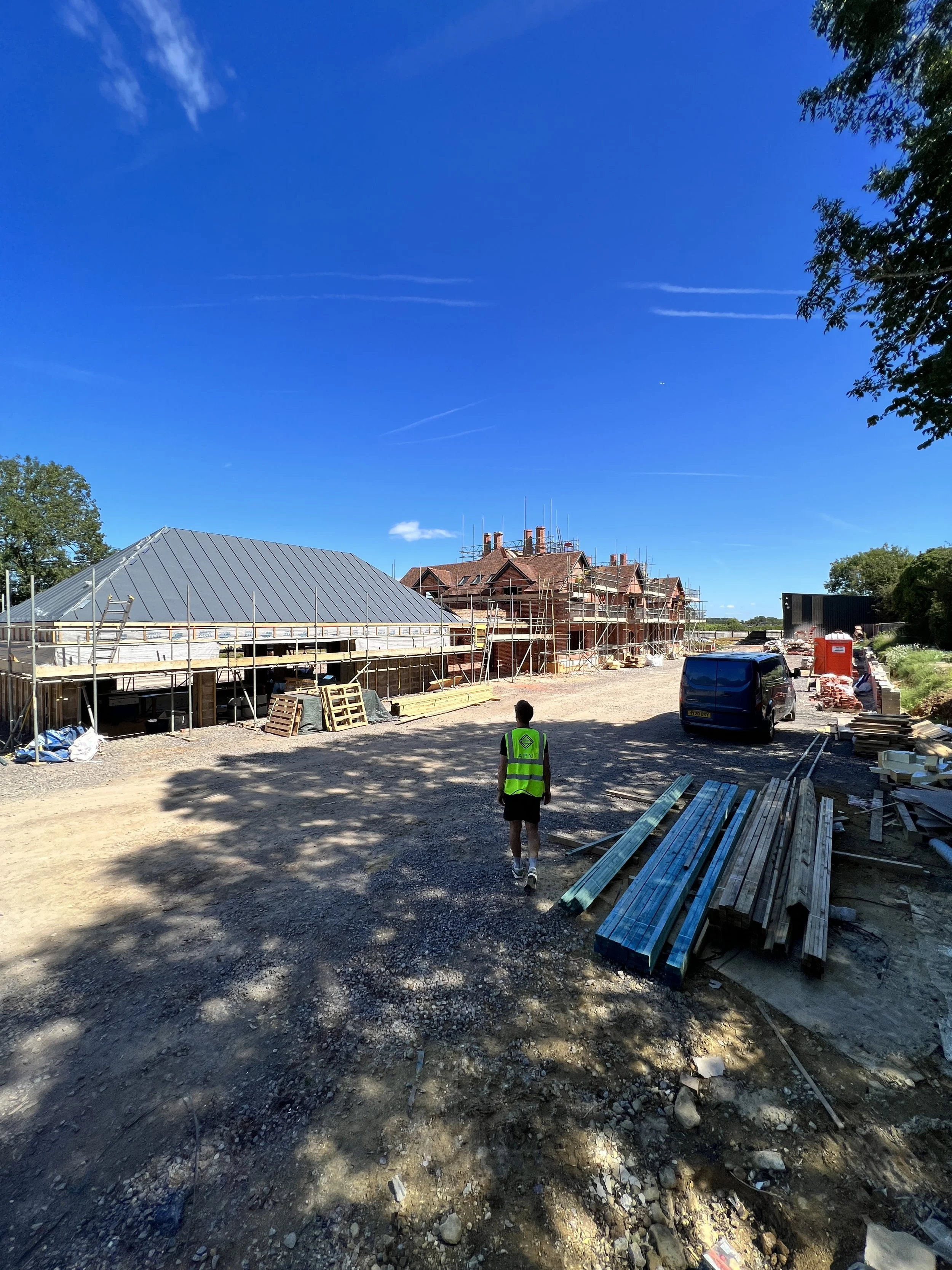 Construction site with a person in a high-visibility vest walking on dirt, scaffolding around unfinished buildings, and construction materials and vehicles nearby under a bright blue sky.