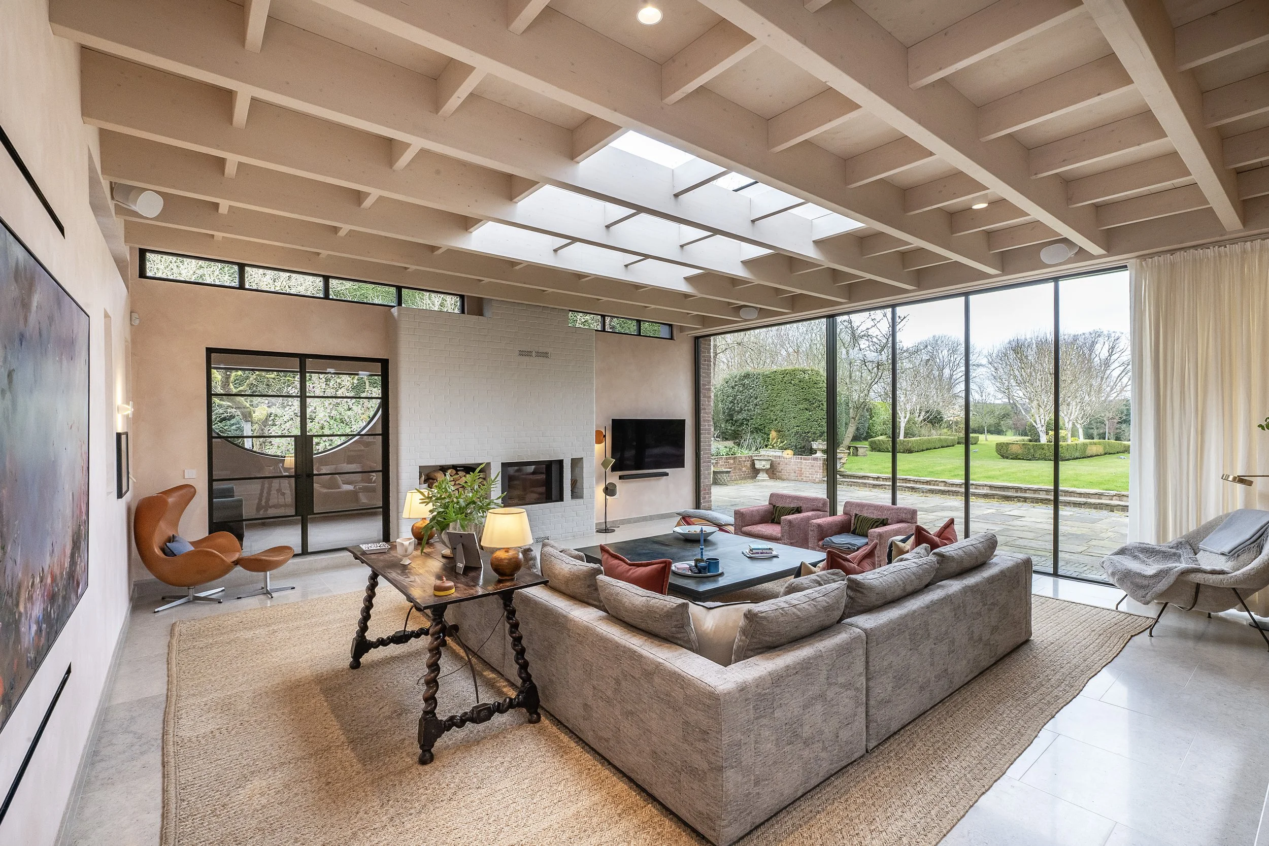 Modern living room with large floor-to-ceiling windows overlooking a lush outdoor garden, featuring a spacious gray sectional sofa, a black coffee table, two orange armchairs, and various decorative items and lamps.