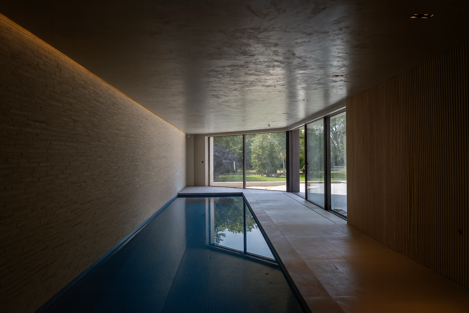 Indoor pool area with large windows, view of trees outside, brick and wooden wall, and tiled flooring.