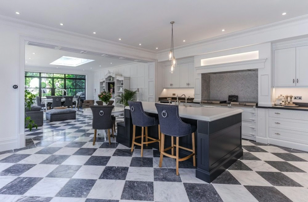 Modern open-concept kitchen with black and white checkered tile floor, white cabinetry, and a large kitchen island with three blue bar stools. The background features a living area with sofas, chairs, and large windows.