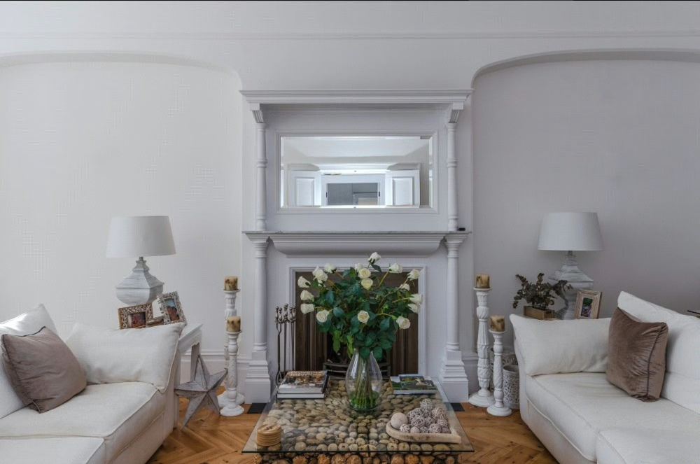 Living room with white sofas, a fireplace with a mirror above, side tables with lamps, and a glass coffee table with a vase of white roses.