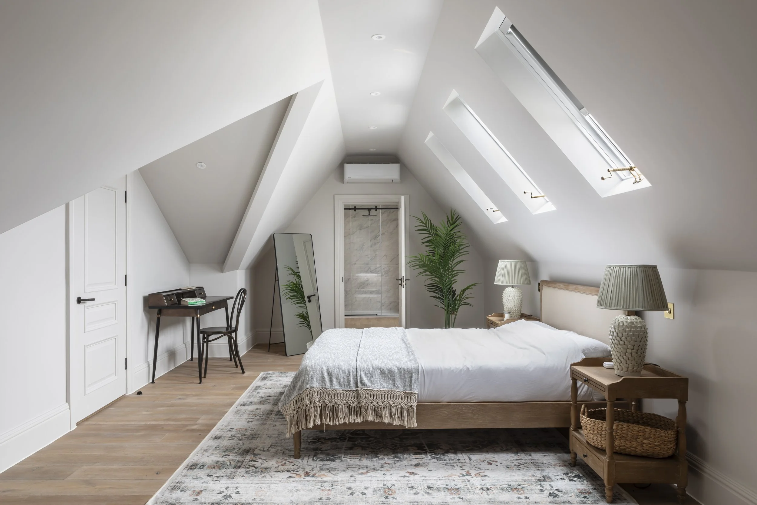 Modern bedroom with sloped ceiling, skylight windows, and minimal decor, featuring a bed with white bedding, bedside lamps, potted plants, a mirror, and a small writing desk.