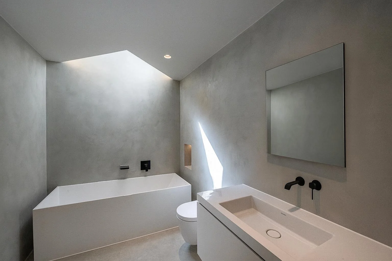 Modern minimalist bathroom with a built-in bathtub, a wall-mounted sink with a large mirror, and a small skylight with natural light.