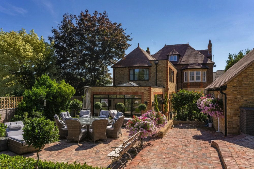 A backyard patio with outdoor seating, surrounded by lush greenery and flowering bushes, and a large historic brick house in the background.
