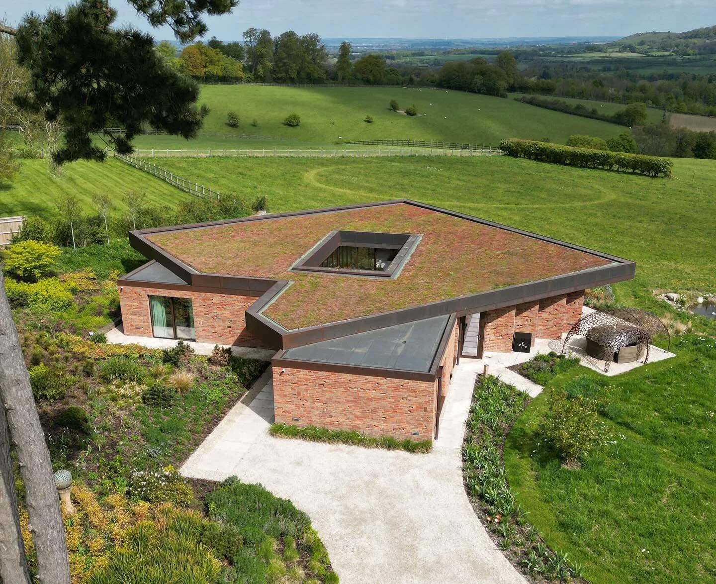 A modern house with a brick exterior and unique angular rooflines in a lush green landscape with open fields and trees in the distance.