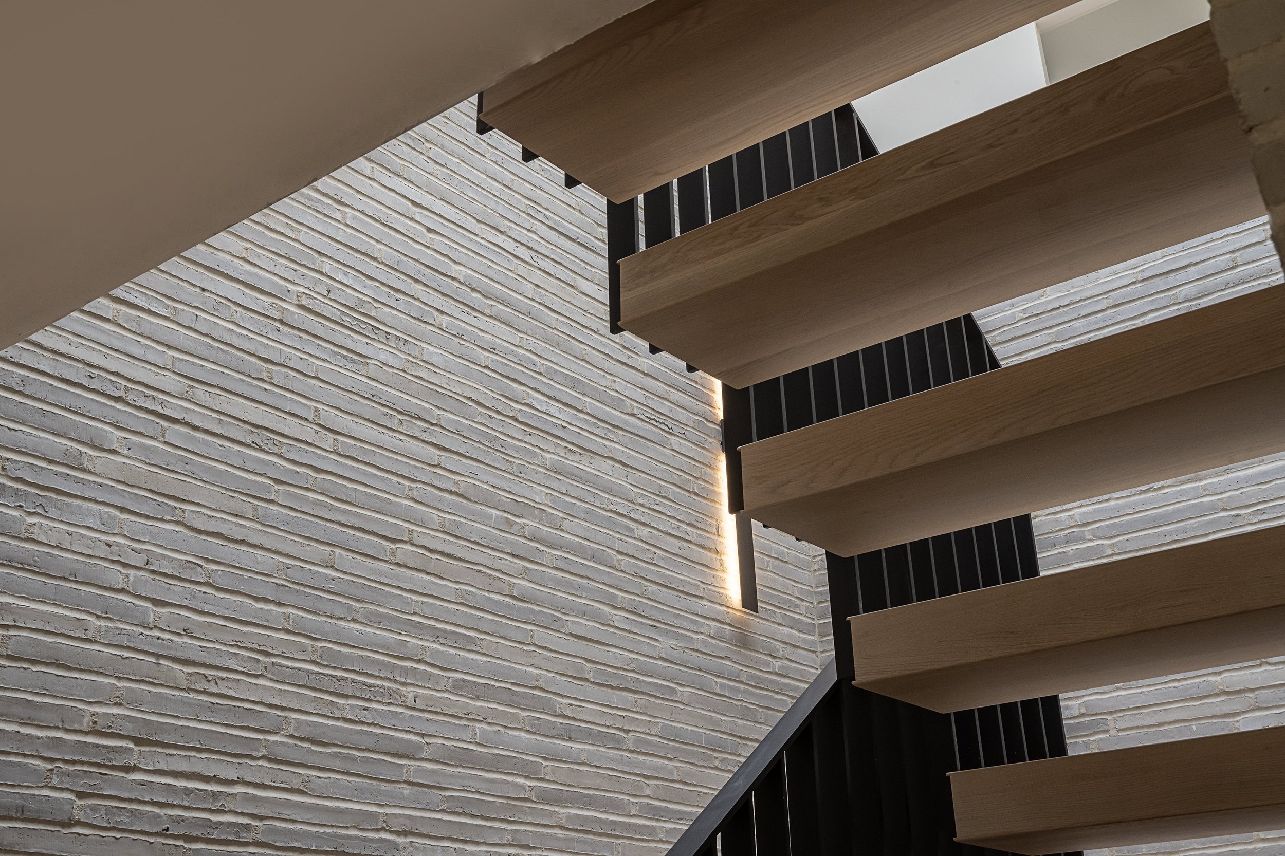 Close-up view of a modern staircase with wooden steps and black railings against a white brick wall with vertical lighting.