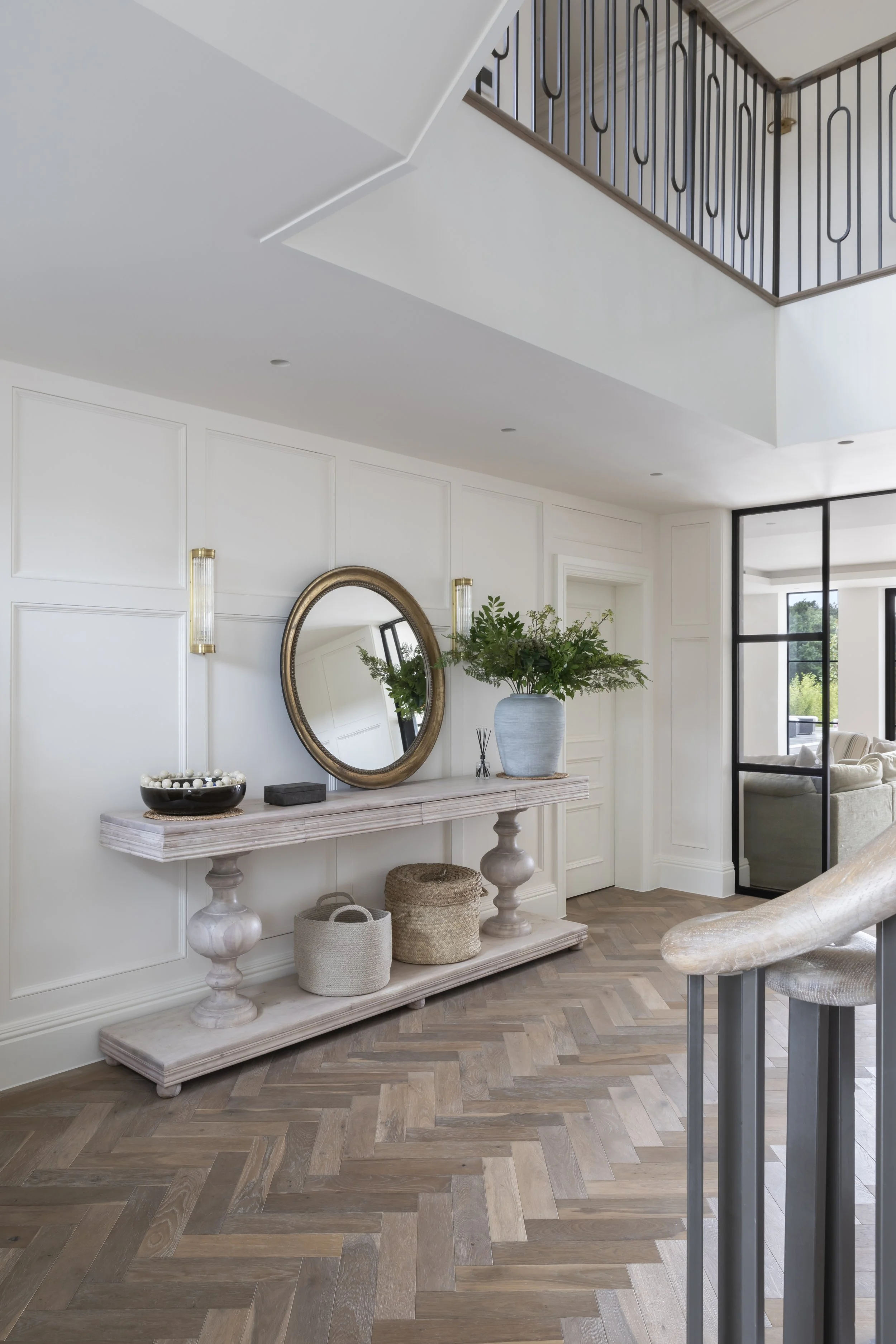 Modern interior entryway with white paneled walls, a wooden console table with decorative items, a large potted plant, and a round mirror hanging above. Light hardwood herringbone floor, with a staircase railing top left and a living room visible in 