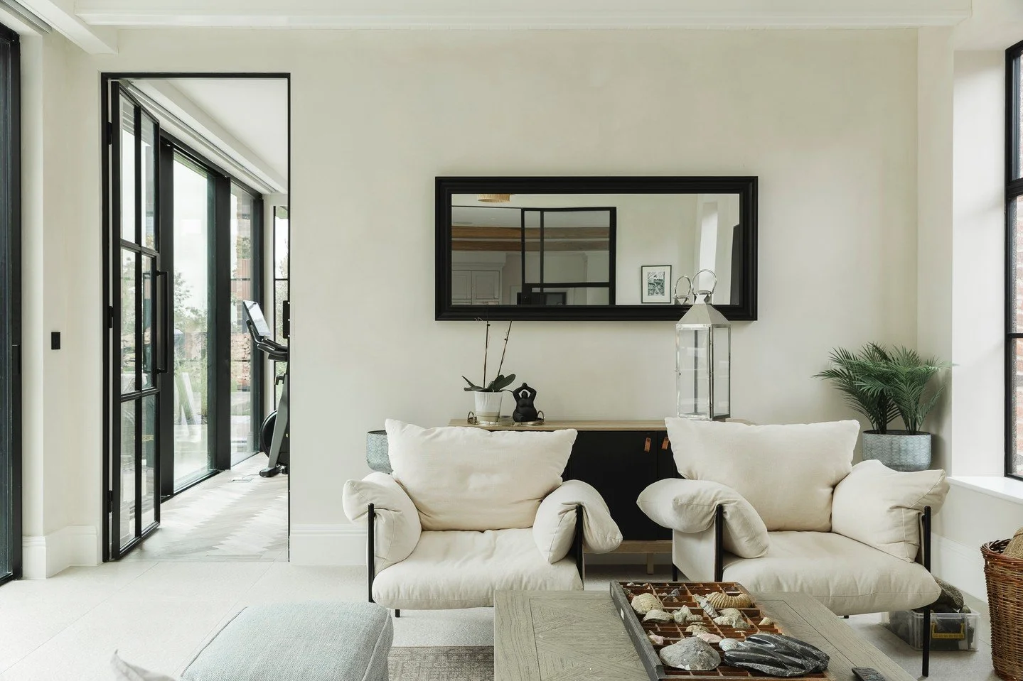 Modern living room with white sofas, black-framed mirror, glass lanterns, potted plant, and sliding glass door leading to outdoor area with exercise equipment.
