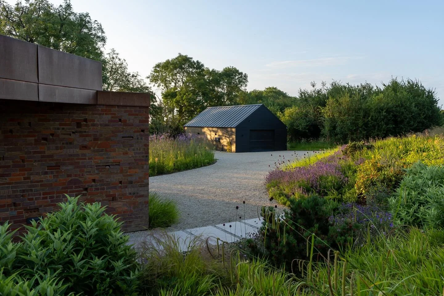A gravel pathway curves through a lush garden with purple and green plants leading to a black shed with a metal roof in the distance. A brick wall is visible on the left side of the image.