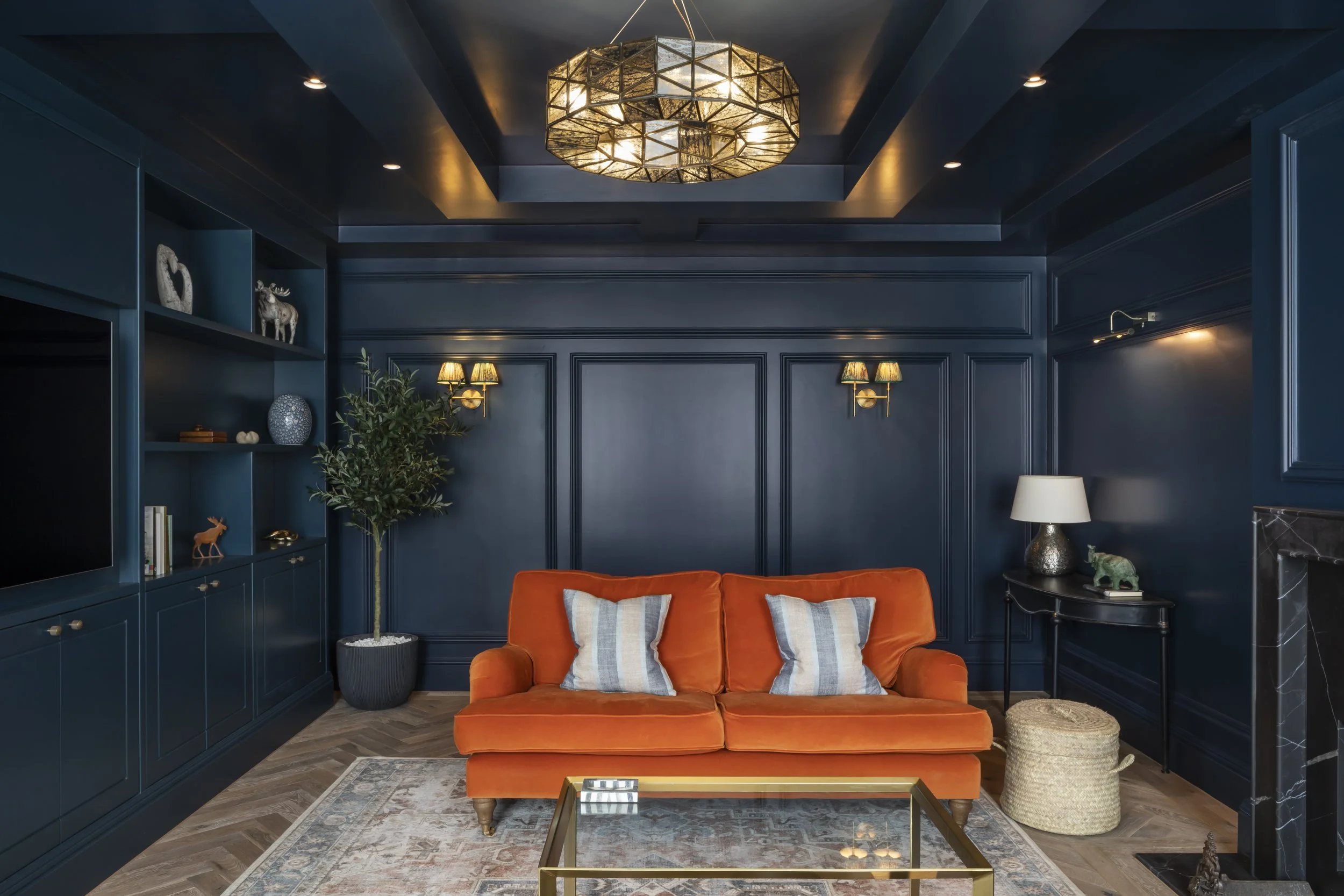 Living room with navy blue walls, orange velvet sofa with striped pillows, green potted plant, black side table with lamps, and a glass coffee table on a patterned rug.