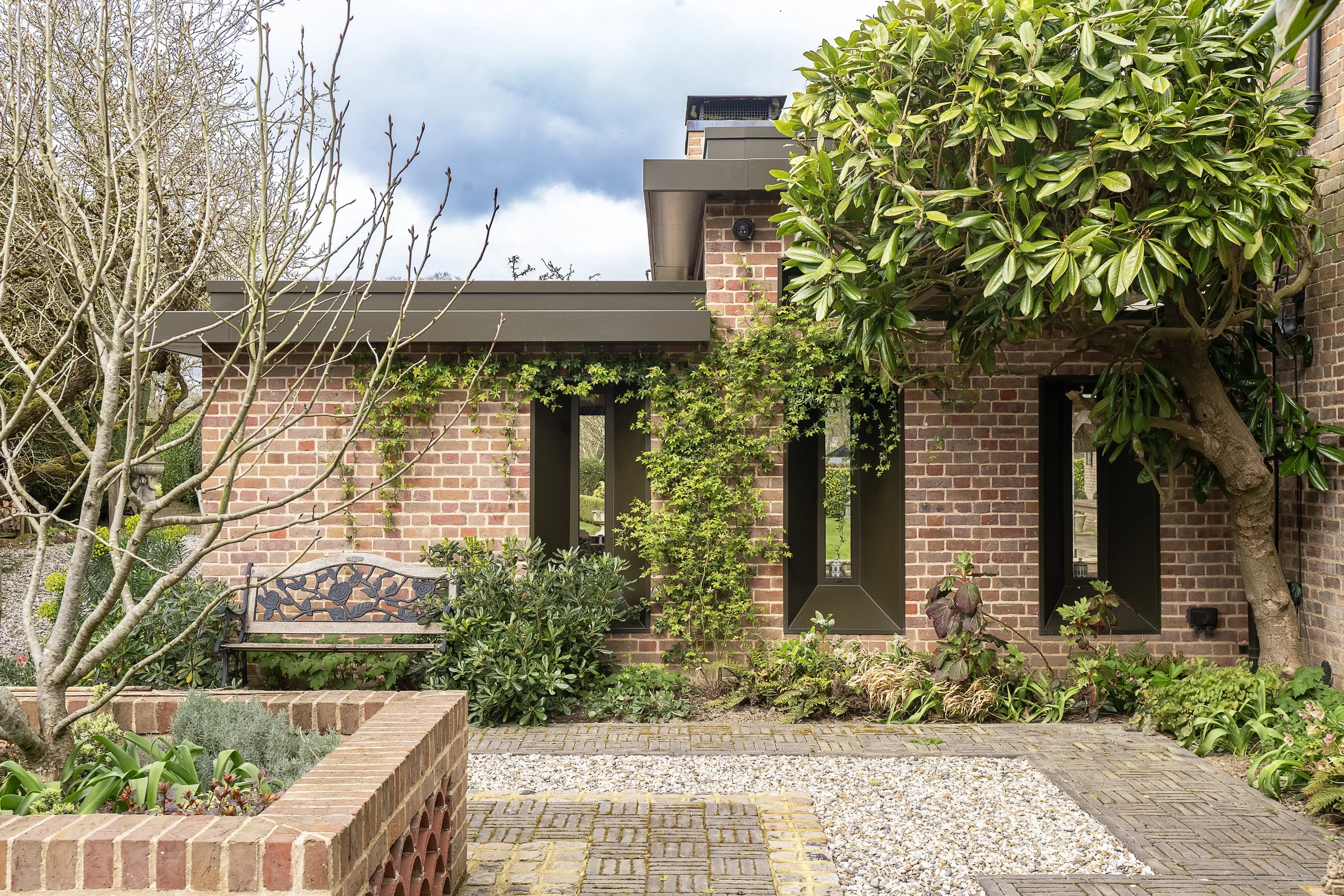 Front view of a brick house with modern black-framed windows, surrounded by a garden with various trees, shrubs, and a stone and brick pathway.