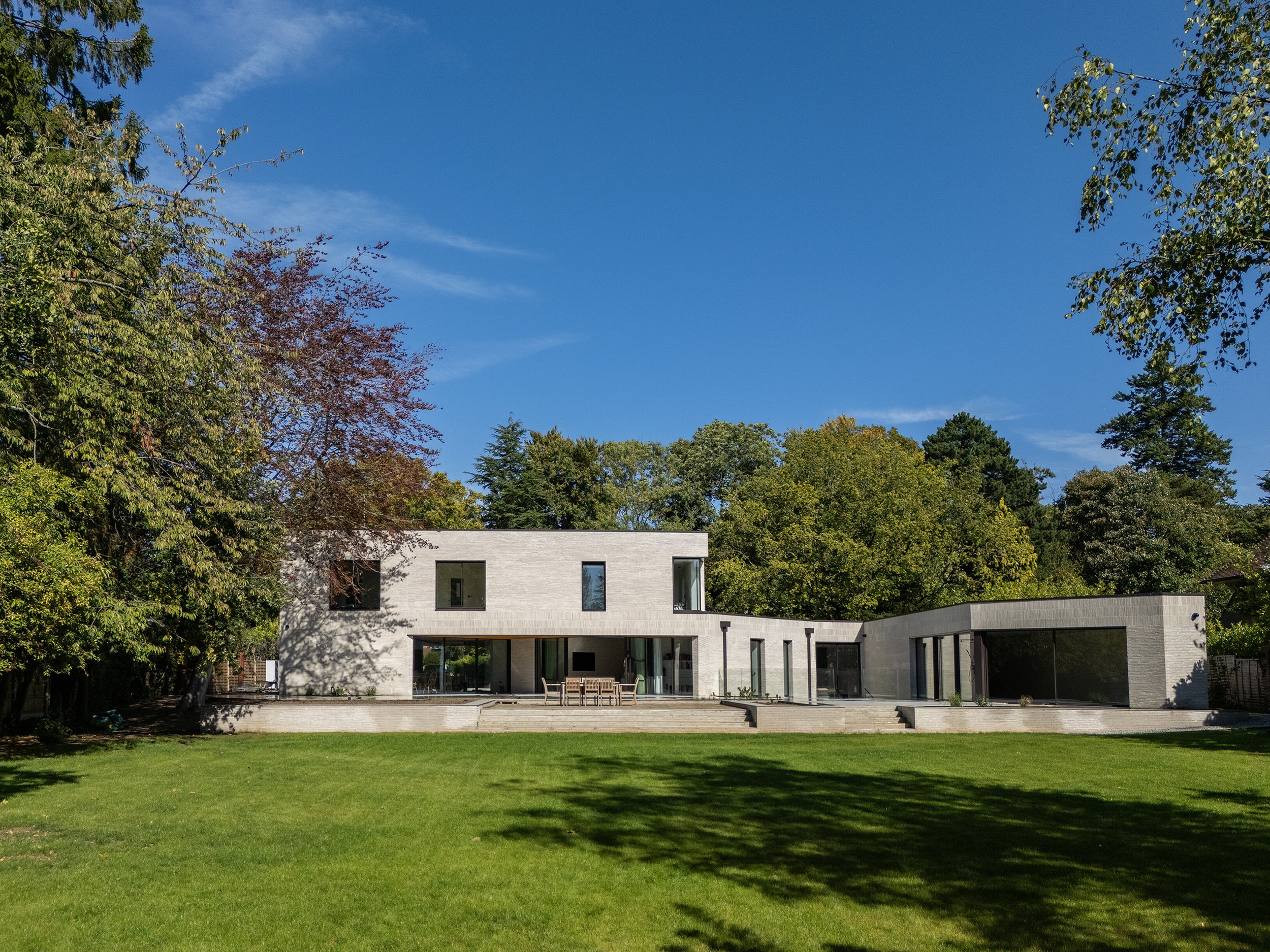 Modern white house with large glass windows and a flat roof, surrounded by green trees and a grassy lawn under a blue sky.